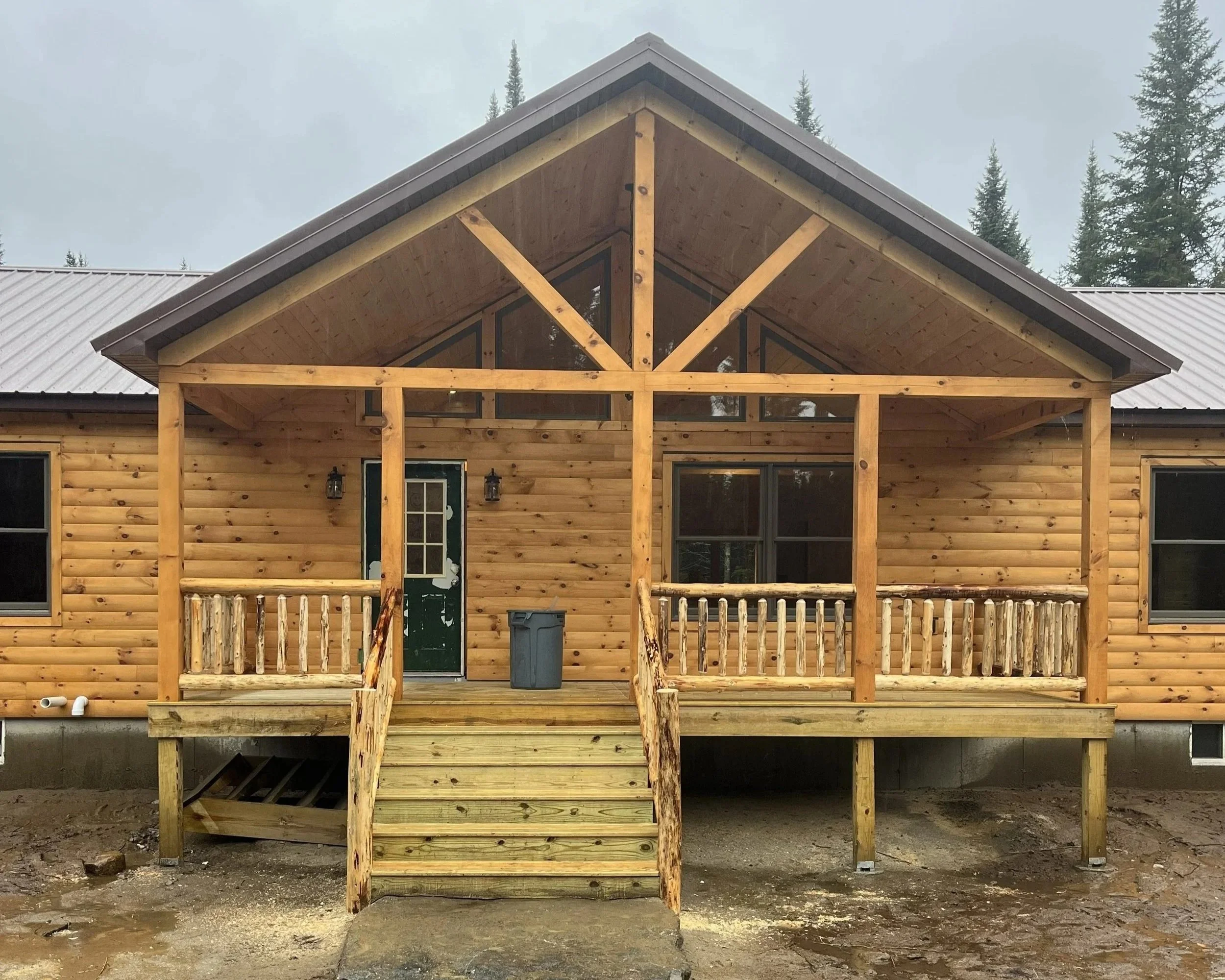 Front view of a wooden house under construction with a porch, surrounded by trees and cloudy sky.