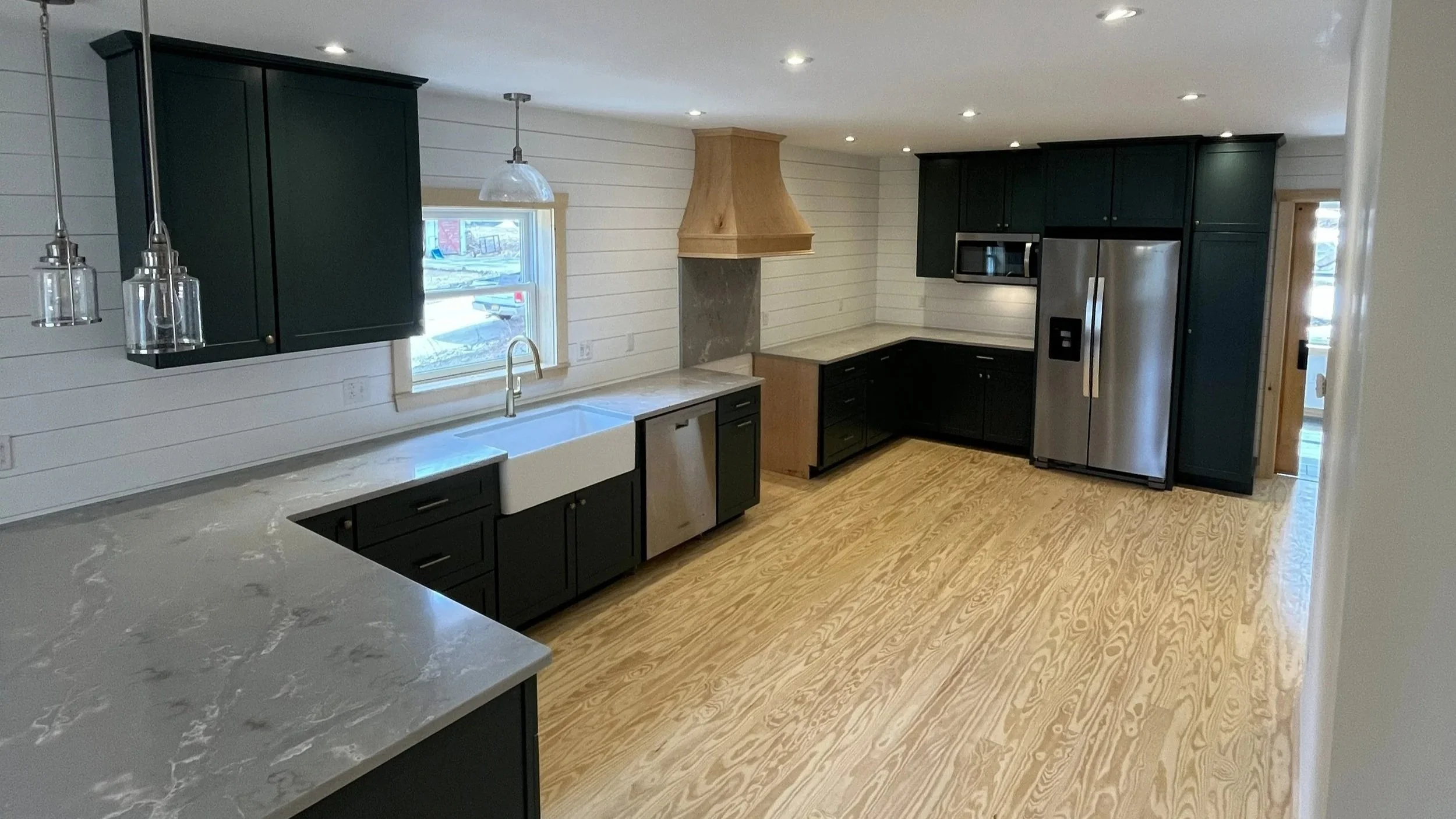 Modern kitchen with black cabinets, stainless steel refrigerator, microwave, white farmhouse sink, wooden range hood, and light wood flooring.