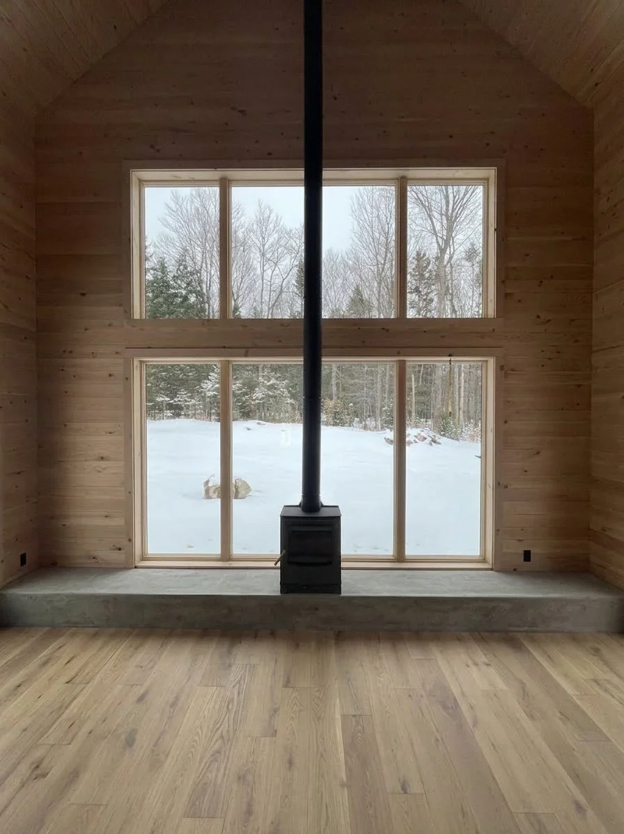 Interior view of a room with large windows, wooden walls, and a wood stove located on a concrete platform, with a snowy outdoor landscape visible through the windows.
