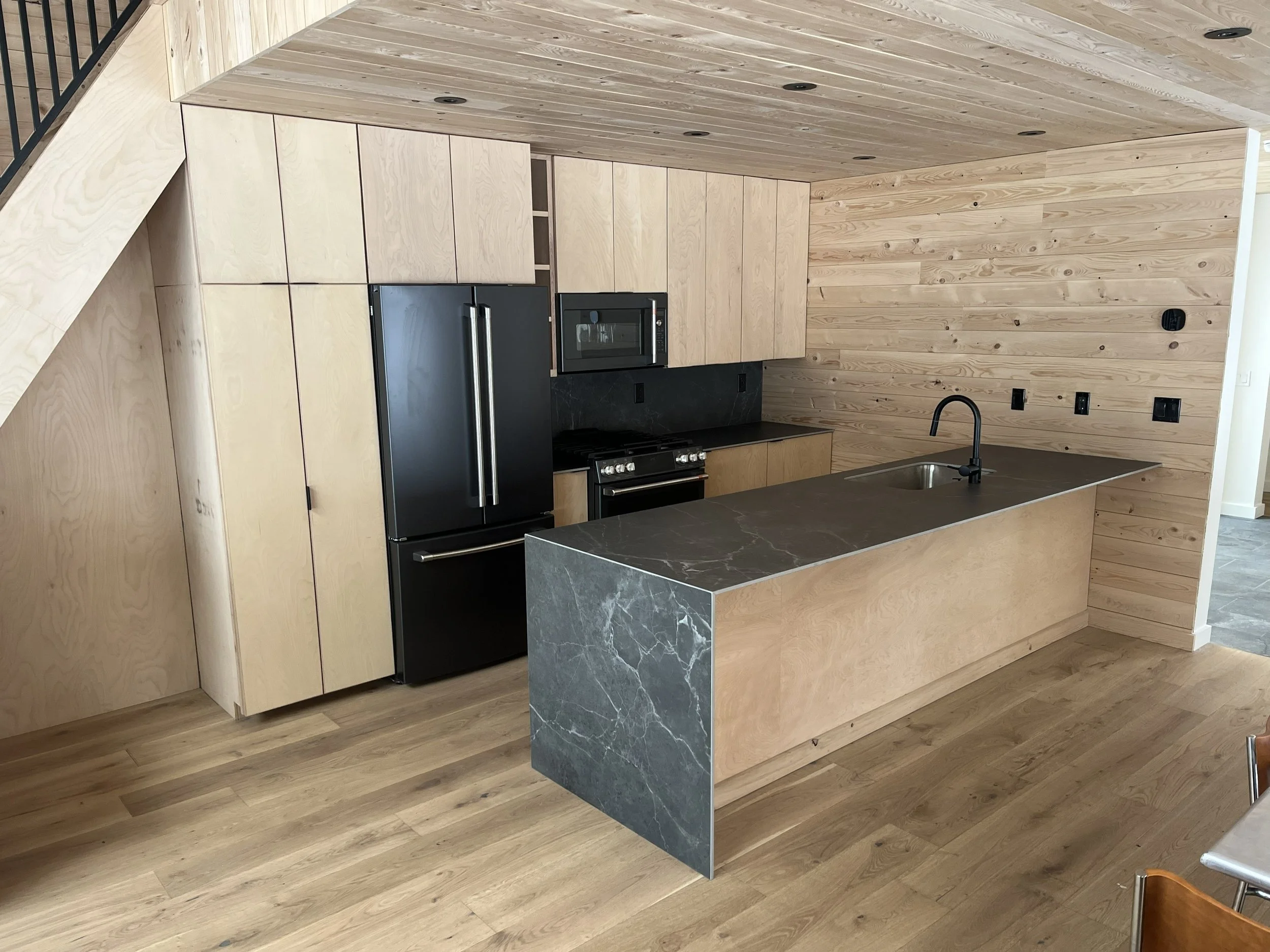 Modern kitchen with light wood cabinets, black appliances, a dark marble island, and wood-paneled walls and ceiling.