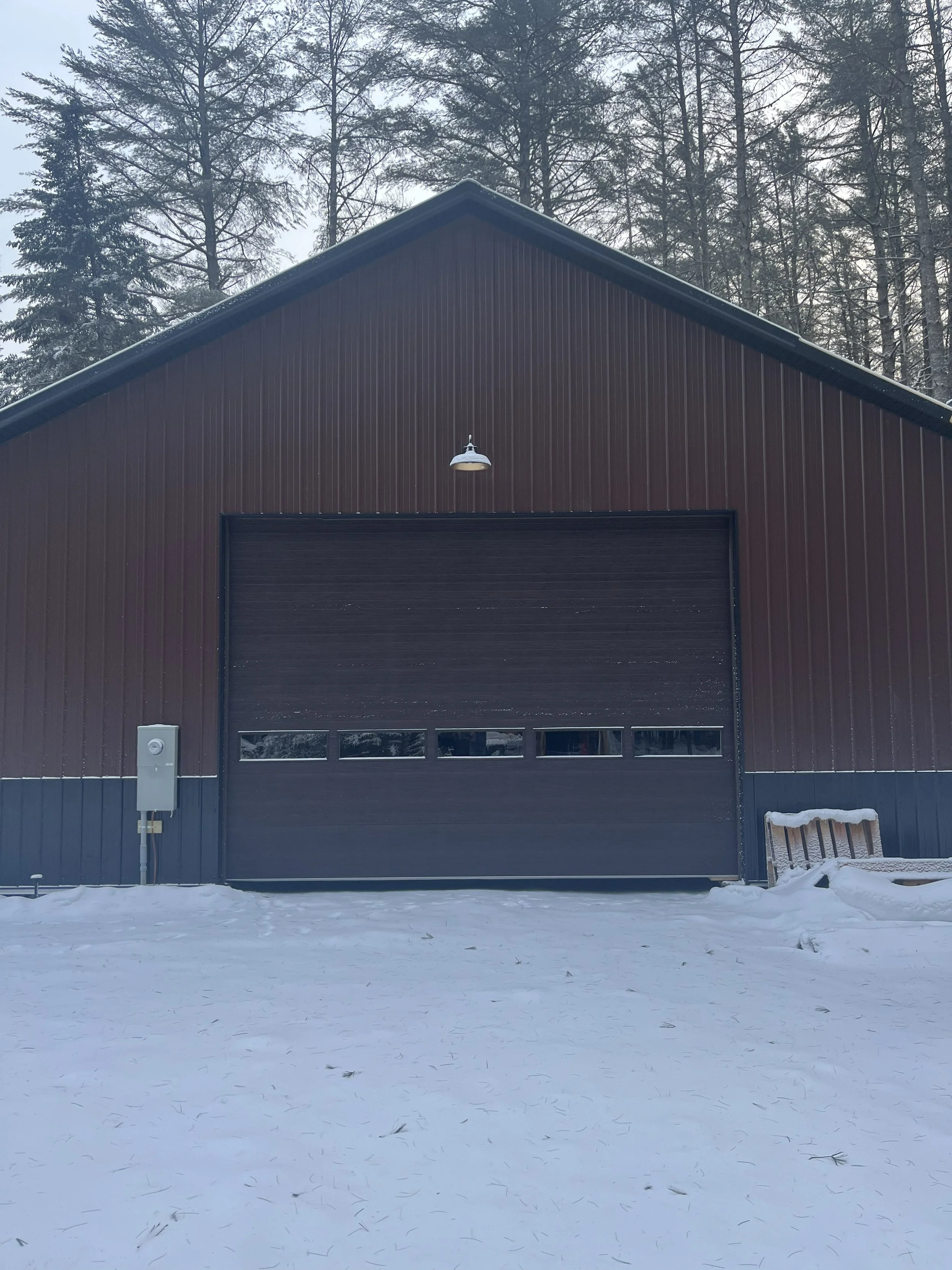 A dark brown garage with a large roll-up door, set outdoors in winter, with snow on the ground and a background of tall, leafless trees and a cloudy sky.