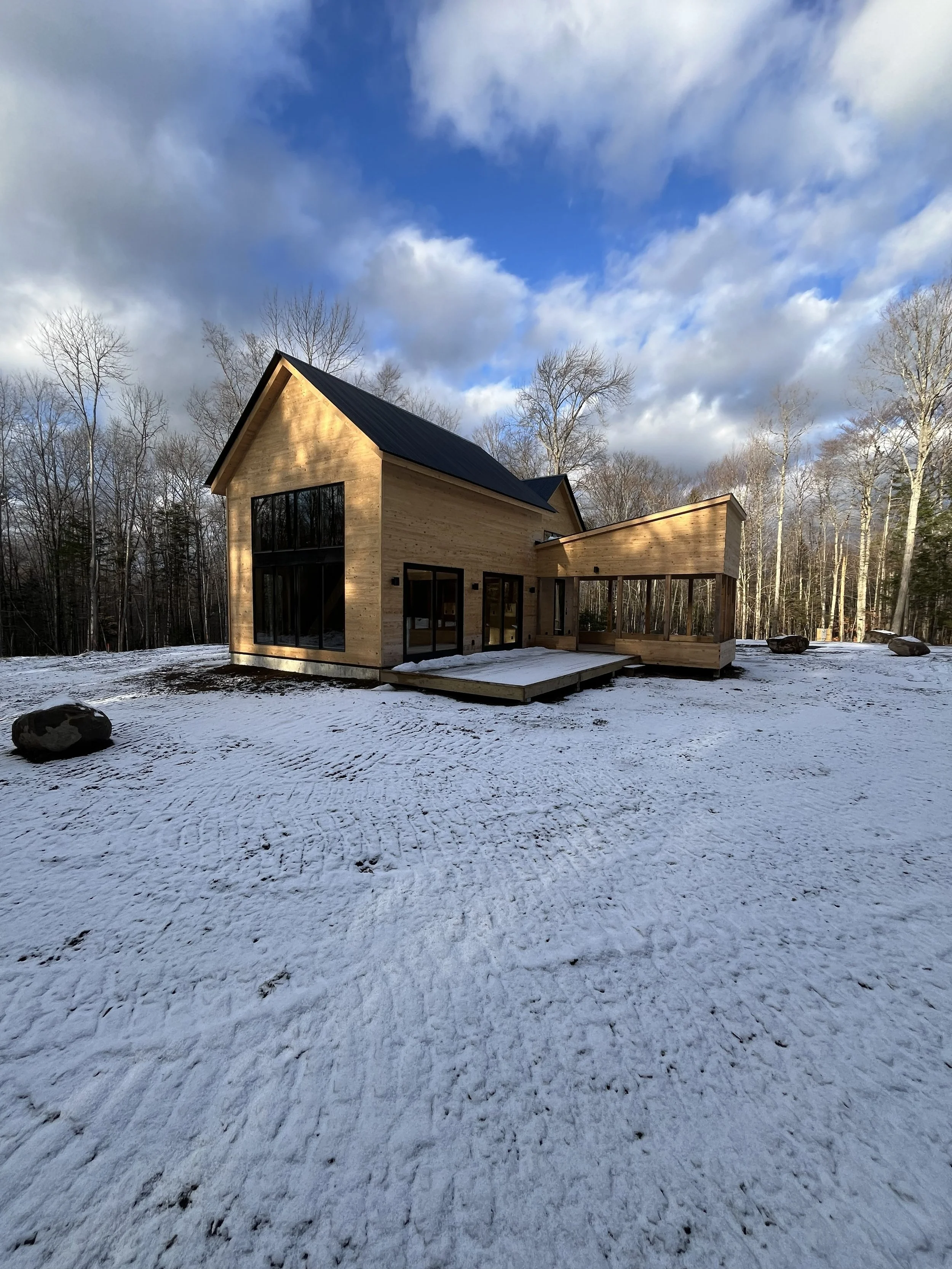A modern wooden house with large windows in a snowy landscape, surrounded by leafless trees under a partly cloudy sky.