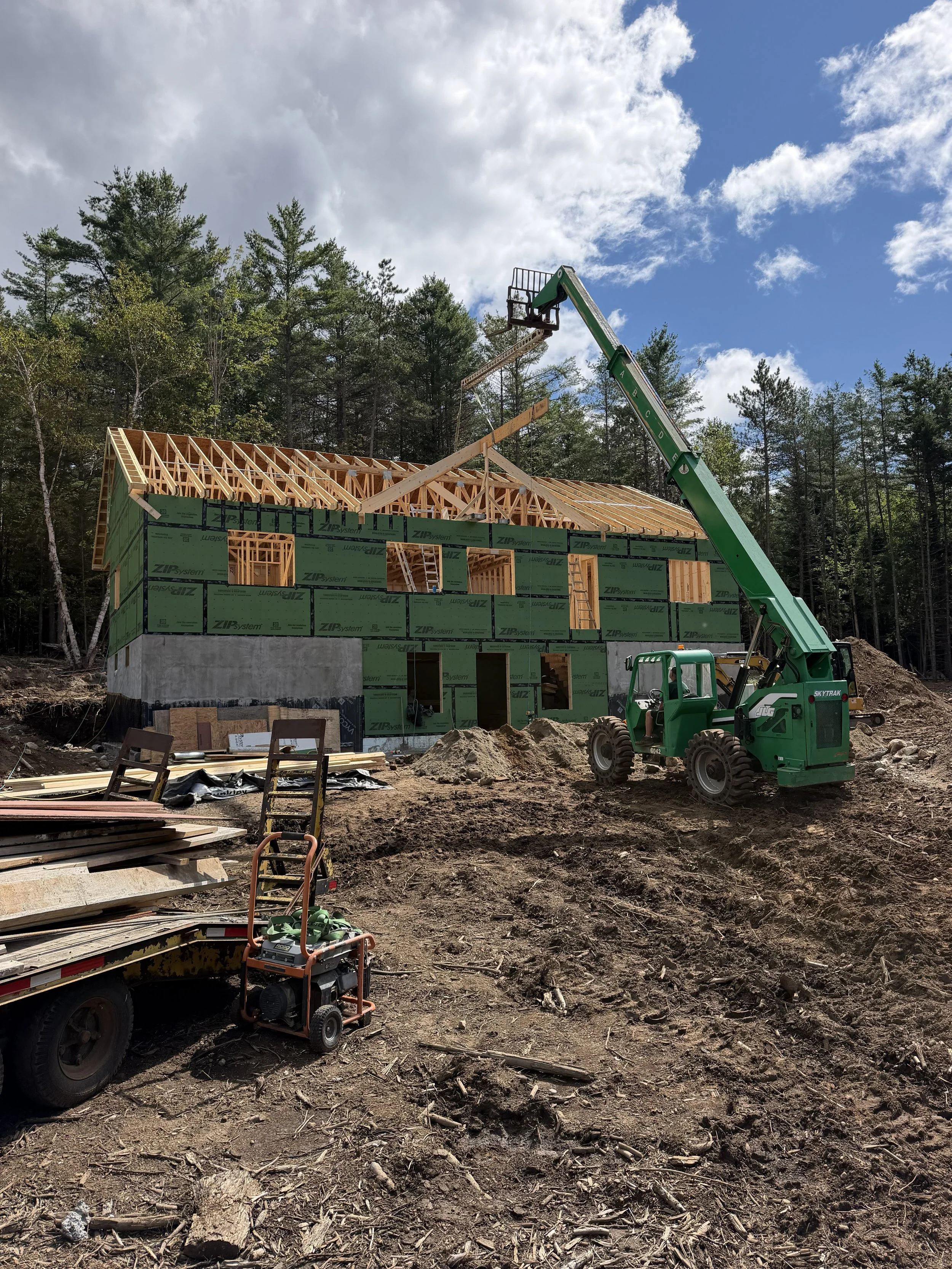 Construction site with a house frame being built, surrounded by trees and cloudy sky, with construction equipment and materials on the ground.