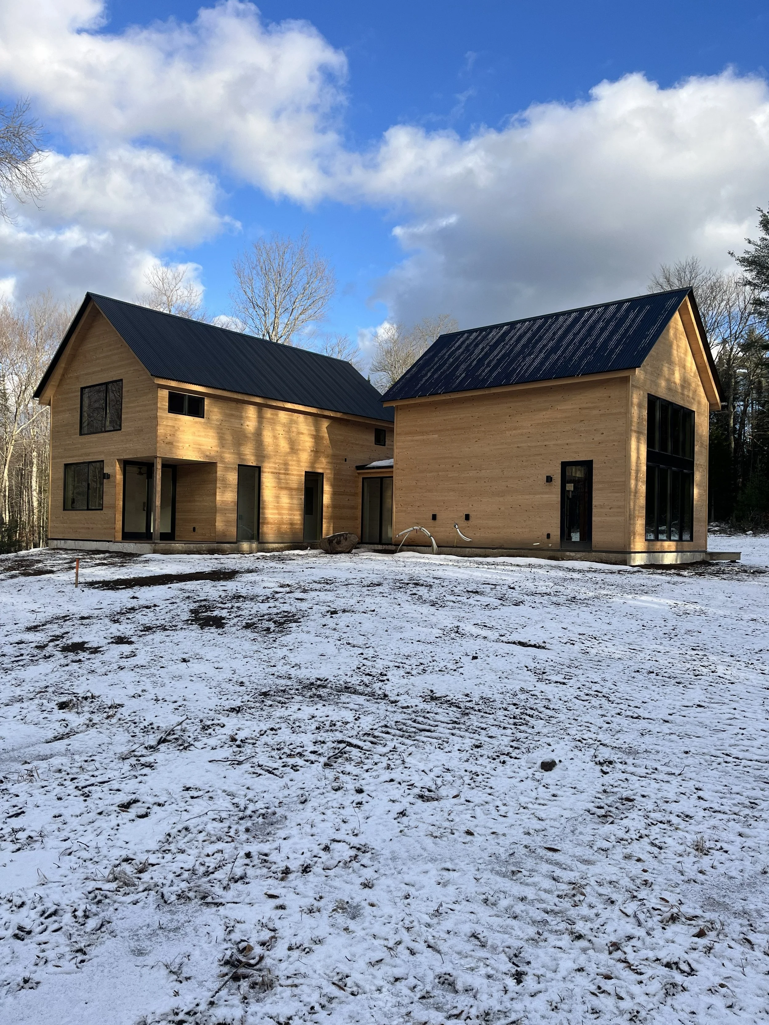 A Scandinavian style house with wooden exteriors and black roofs, surrounded by a snowy landscape with patches of exposed ground and leafless trees under a cloudy blue sky.