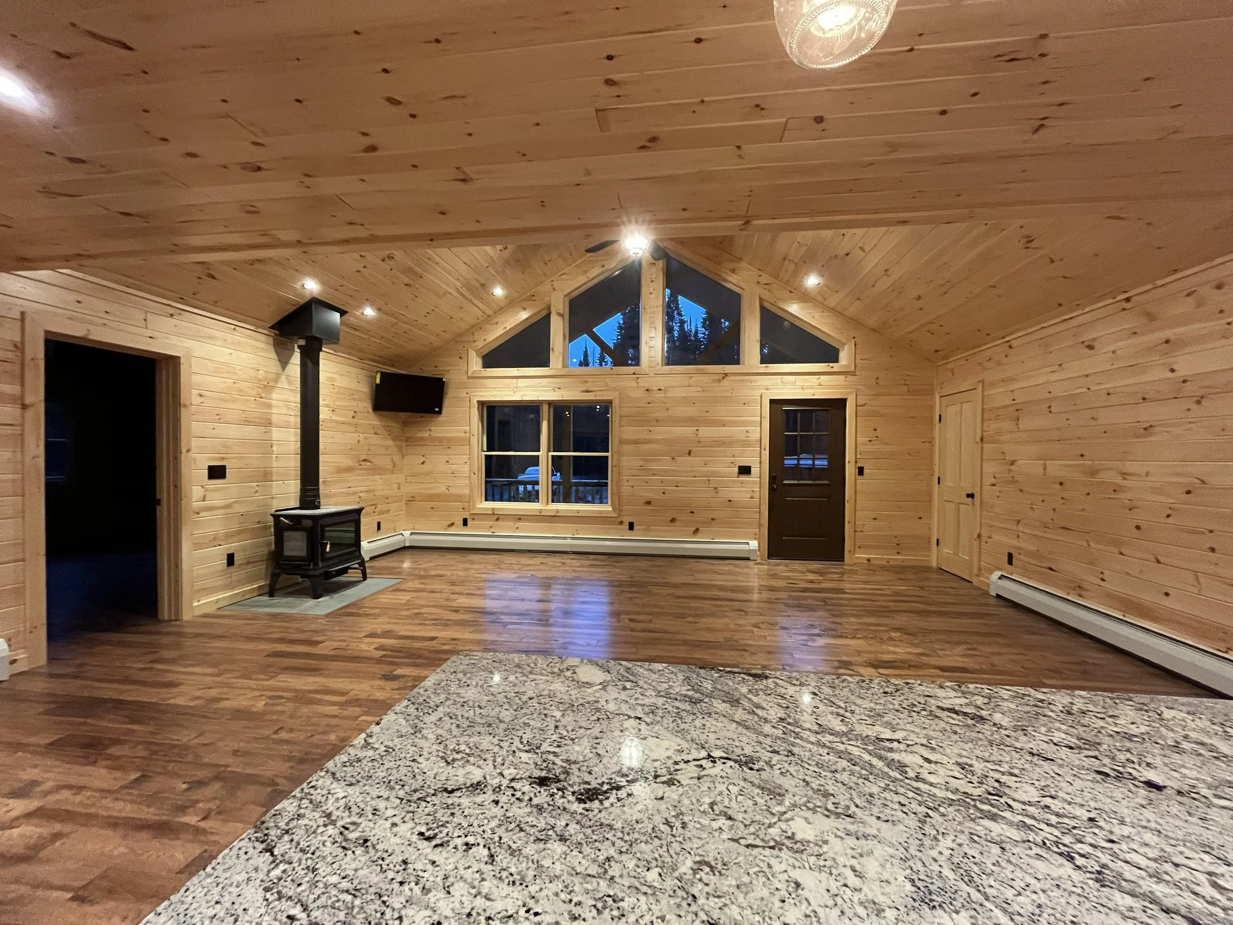 Interior view of a wooden house with high vaulted ceiling, large windows, and hardwood flooring. A wood stove is in the corner, and a flat-screen TV is mounted on the wall. Ceiling lights illuminate the room, and a door leads outside. Mountain trees 