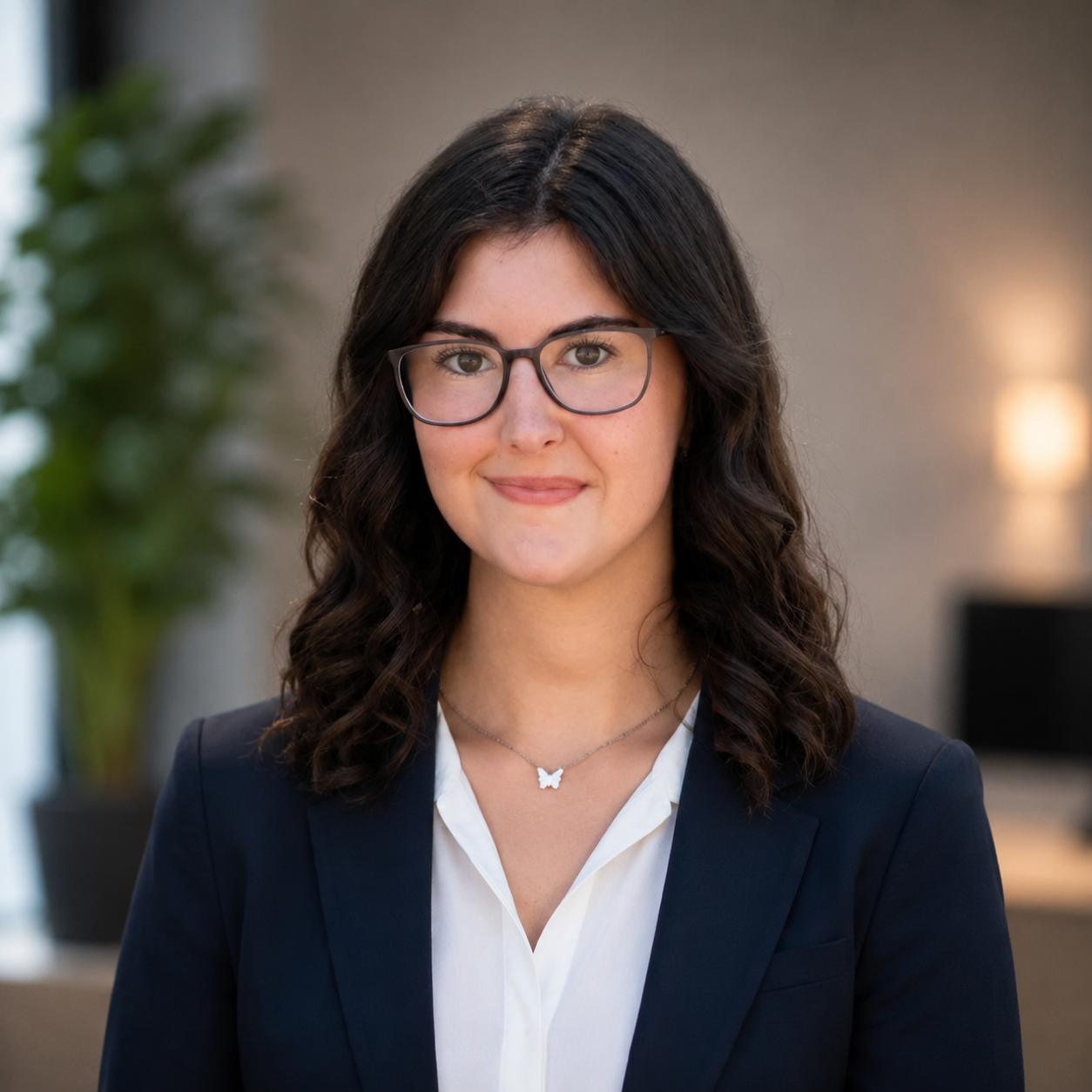 A young woman with dark wavy hair and glasses smiling, wearing a navy blazer and white blouse, in an office setting with a potted plant and soft lighting in the background.