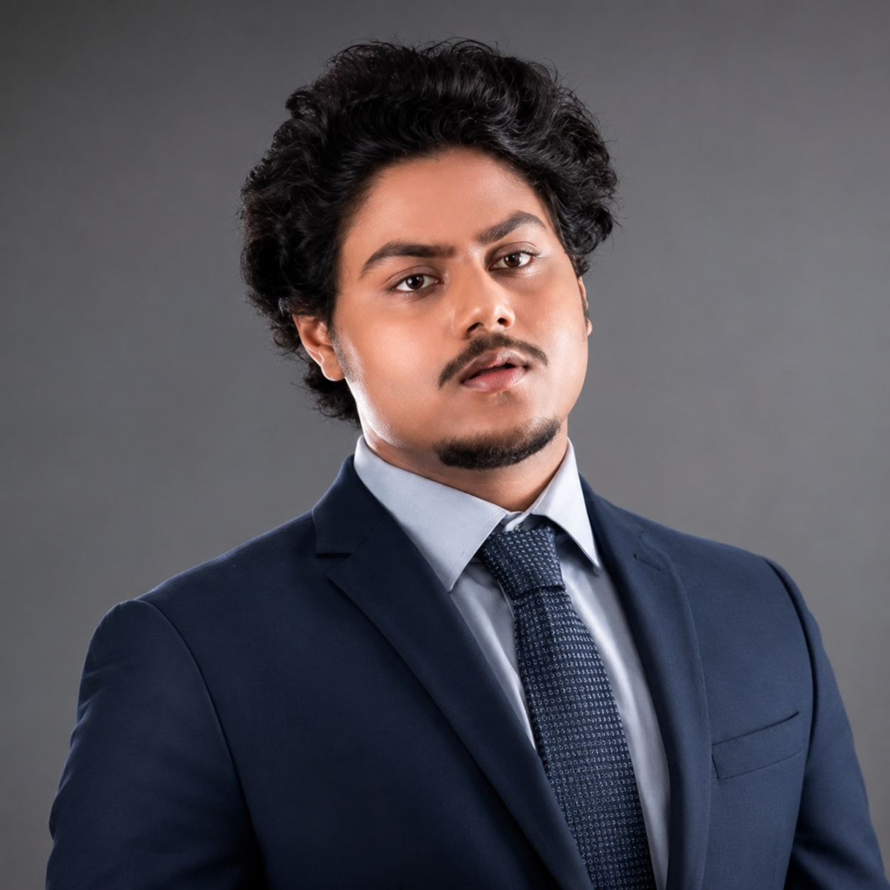 Man with dark curly hair, wearing a navy suit, light blue dress shirt, and navy tie, posing against a gray background.