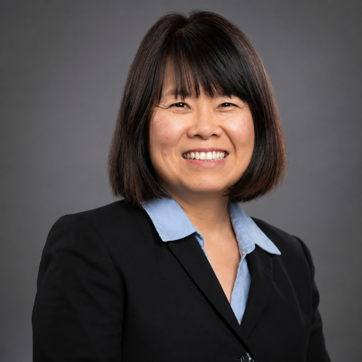 Headshot of an Asian woman with shoulder-length dark hair, smiling, wearing a black blazer over a light blue shirt, against a gray background.