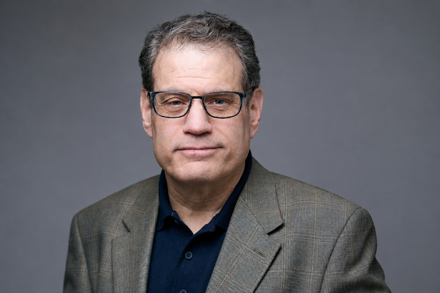 A middle-aged man wearing glasses, a dark collared shirt, and a brown houndstooth blazer against a neutral gray background.