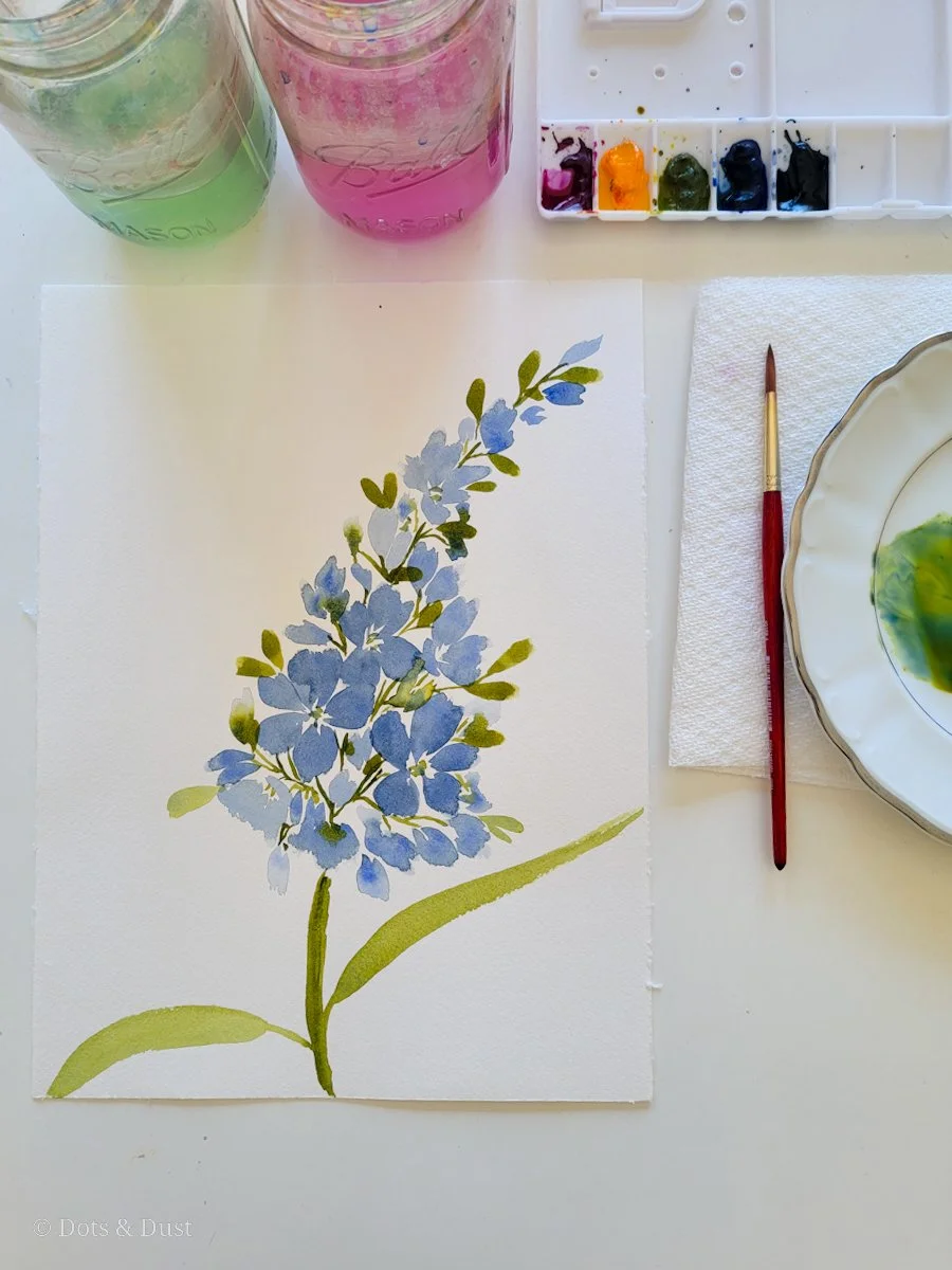 Watercolor painting of blue hydrangea flowers on paper with paint supplies nearby.