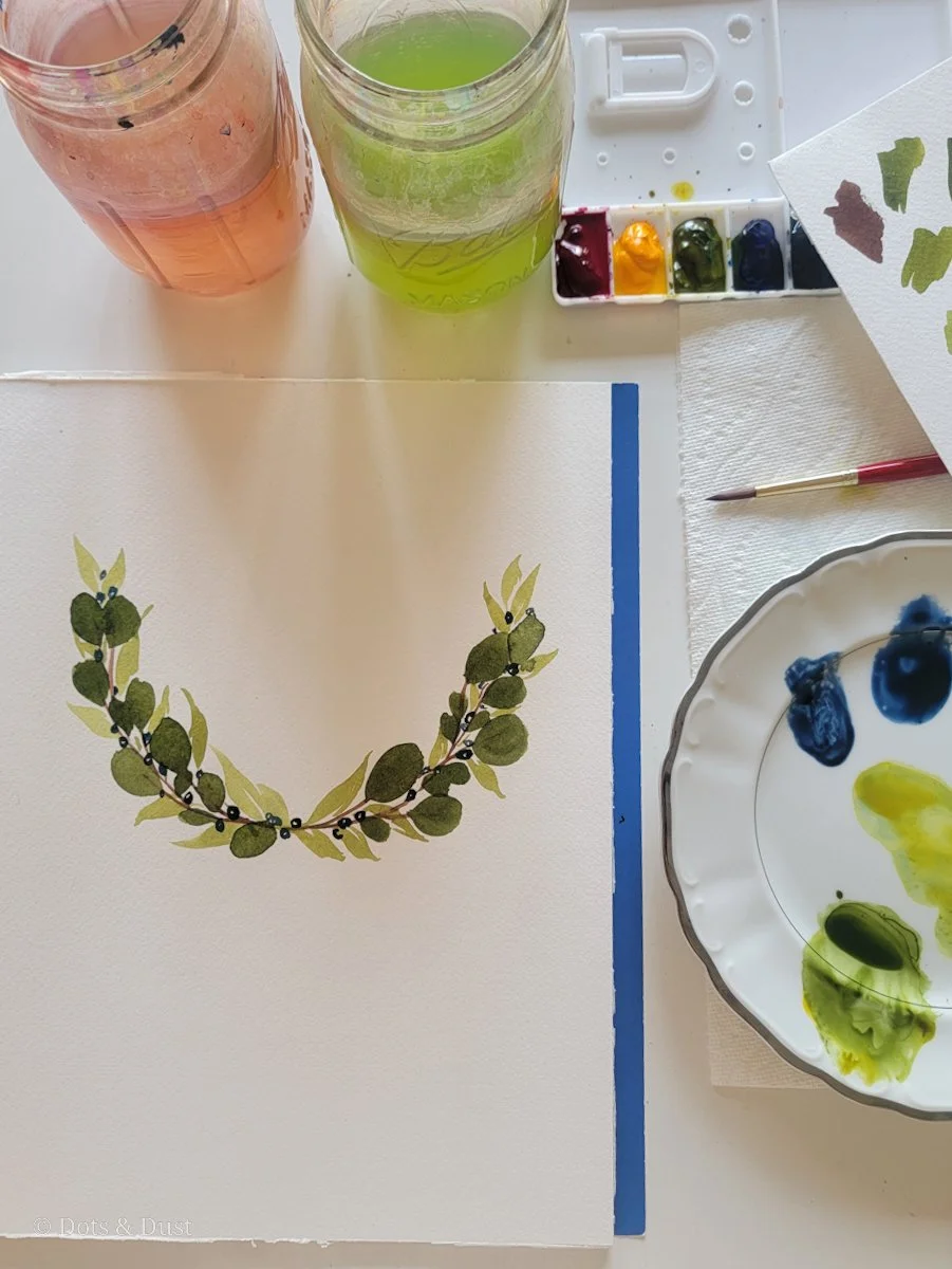 Watercolor painting of a green leafy wreath with black berries, in progress, on a white paper, surrounded by watercolor paints, brushes, and jars of green and pink liquid.