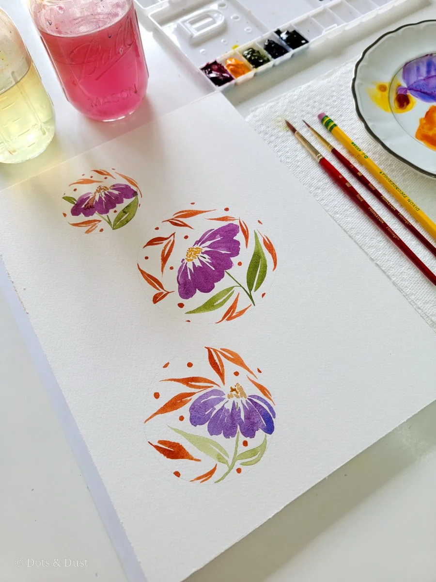 Watercolor painting of three purple flowers with green leaves, surrounded by orange and pink accents, on white paper.