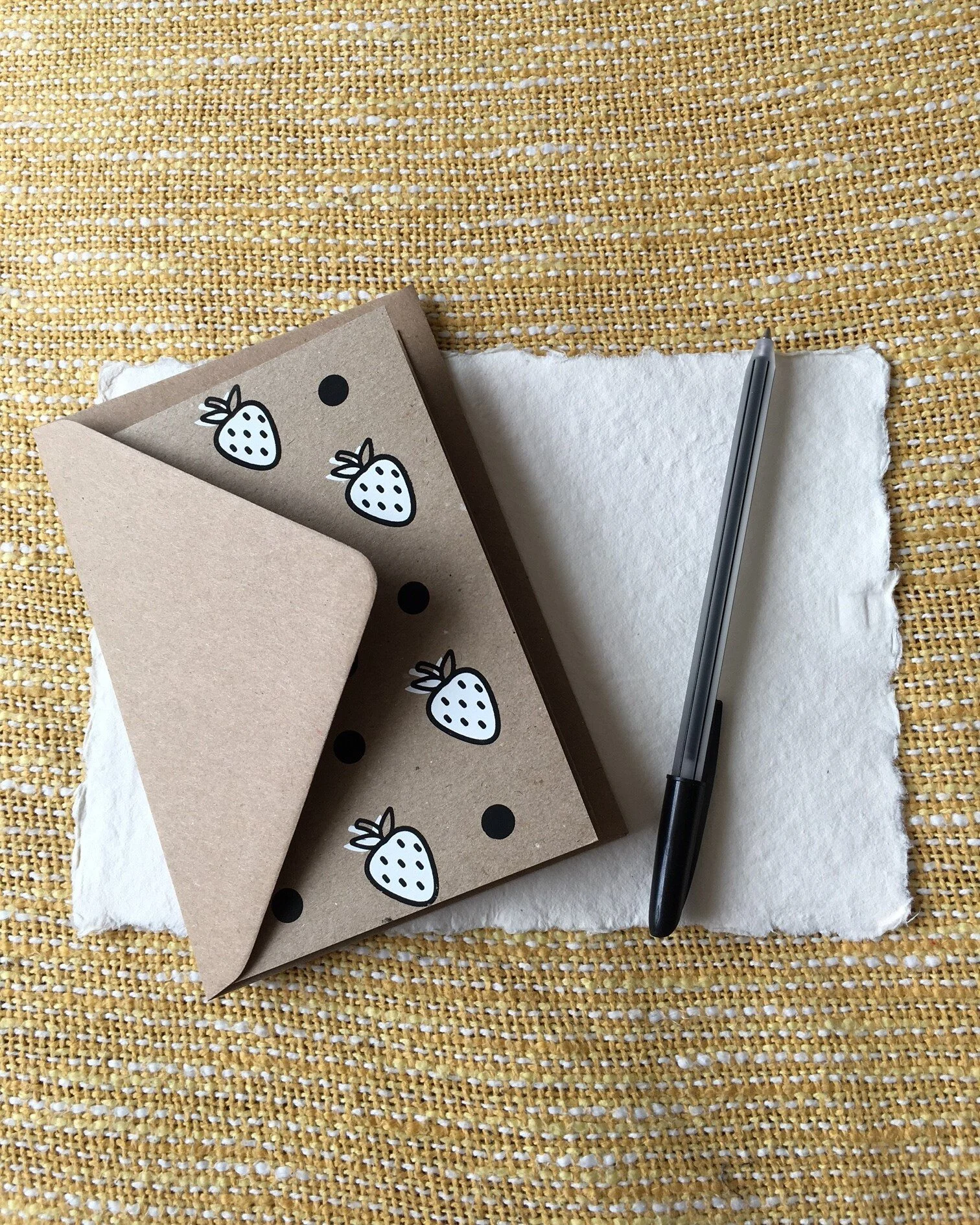 A notecard and pen with white strawberries printed on a plain brown cardstock.
