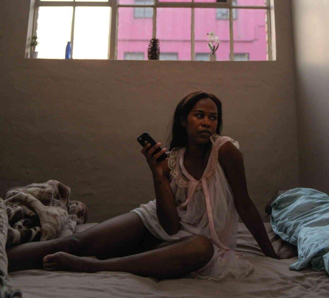  [Image description: A Black woman wearing a pink nightgown sits upright on a bed. She holds a cellphone in her left hand. The sheets of the bed are scrunched at her feet. Through the window above is a pink building in the background. ] 