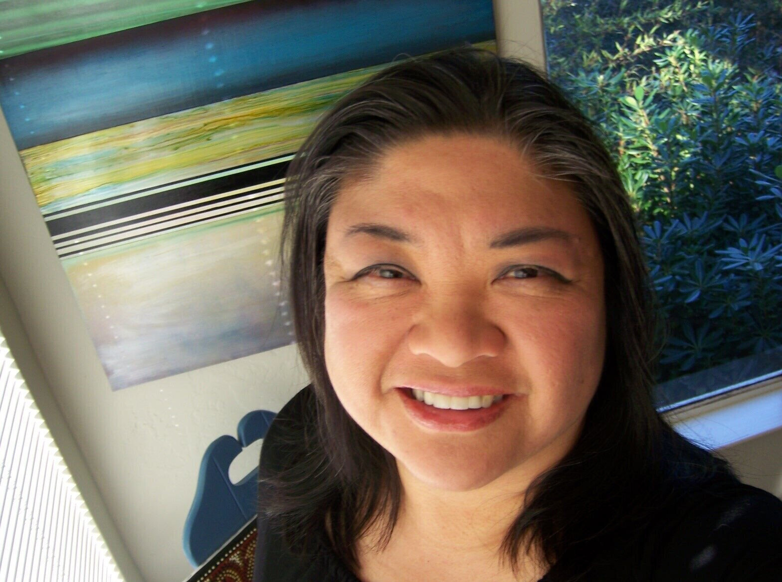  [Image Description: Claire Aguilar is pictured from the shoulders up She stands in front of a screened window and a multicolored abstract painting. She smiles.]  