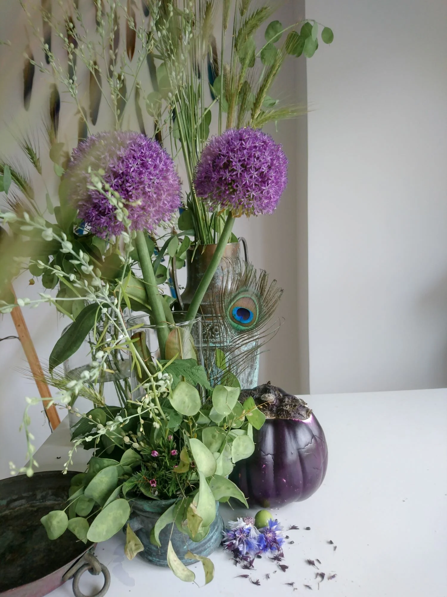 Purple alium flowers sitting on a workbench with a purple eggplant and a peacock feather. Fiori di allium viola sul banco di lavoro con una melanzana e piume di pavone.