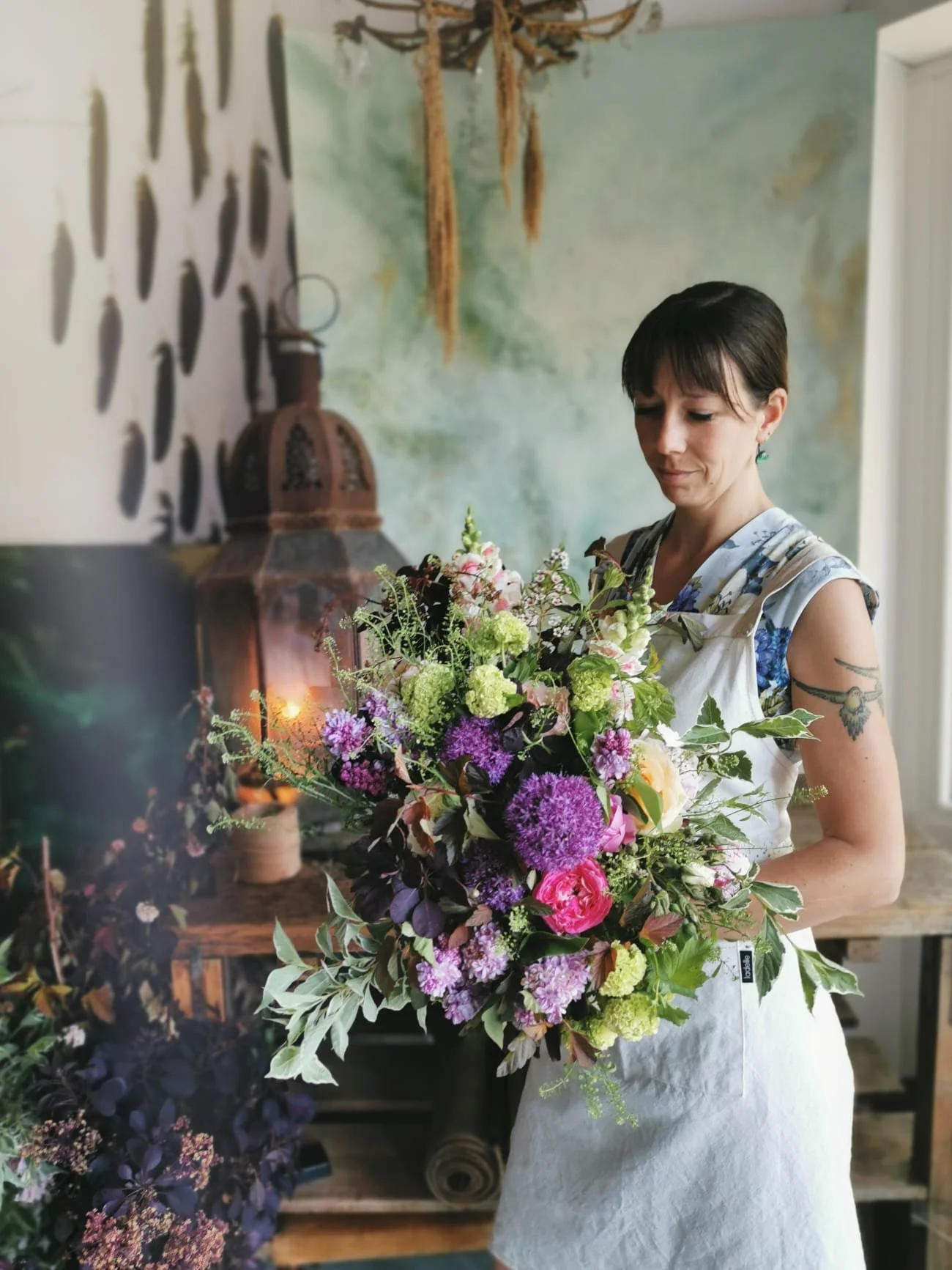 A florist holding a bouquet of seasonal fresh flowers, in a floral design studio next to a glowing rusty lantern with lit candles on an old wooden workbench.