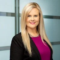 A professional woman with long blonde hair, wearing a black blazer and pink top, smiling in front of a modern office background.