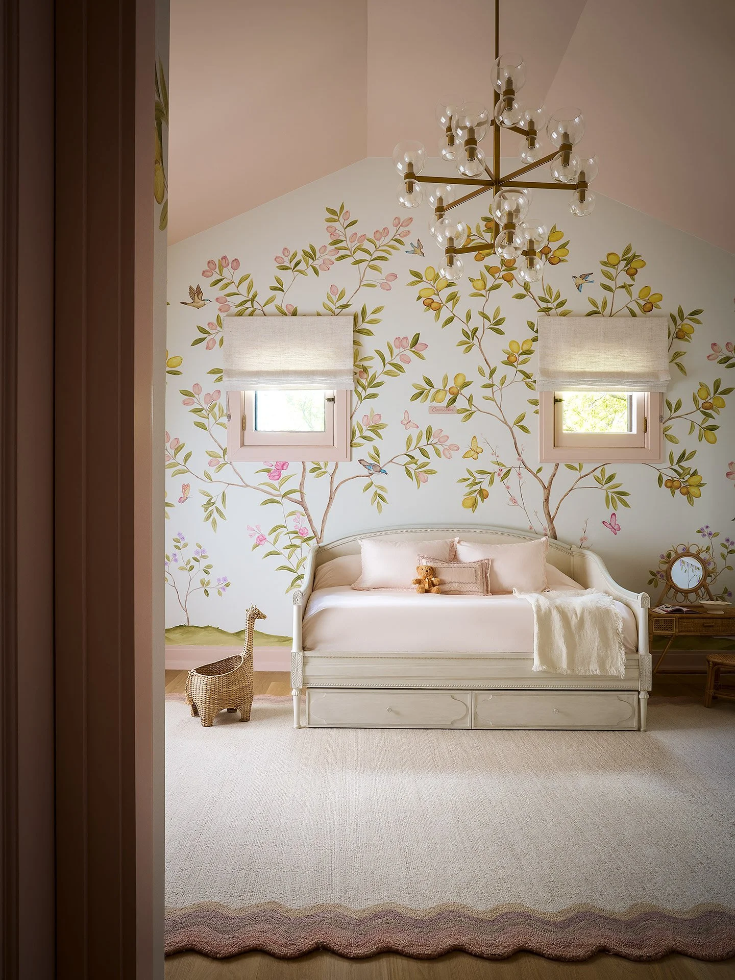 A cozy bedroom with a white wooden bed, pink bedding, a Teddy bear, a wicker giraffe, two small windows with white Roman shades, a chandelier with multiple bulbs, and a mural of trees, birds, and butterflies on the wall behind the bed.