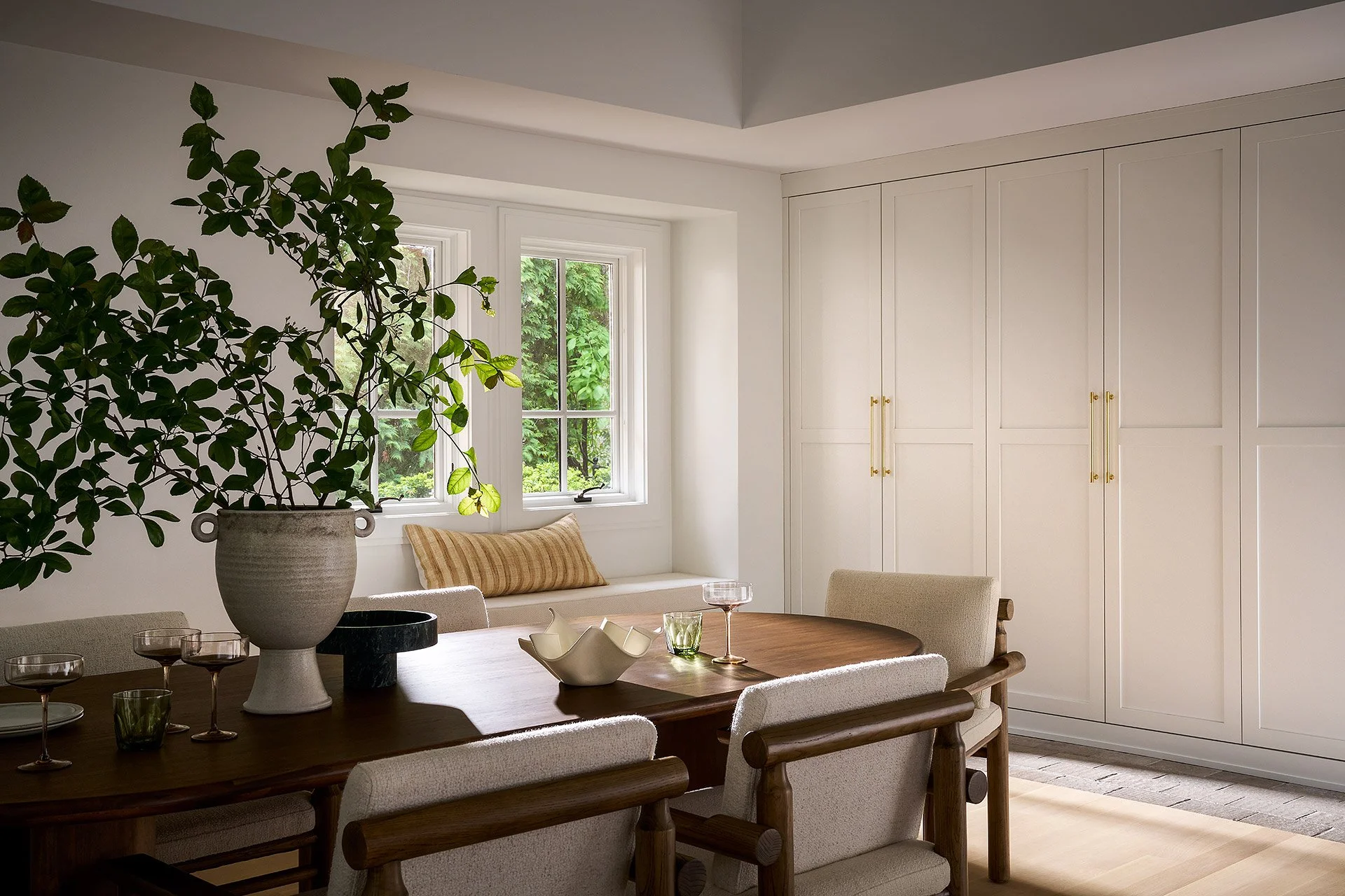 A cozy dining area with a large wooden table surrounded by beige upholstered chairs. On the table are several glasses of wine and a decorative ceramic bowl. A large potted green plant is on the table, and a window with white framing allows natural li