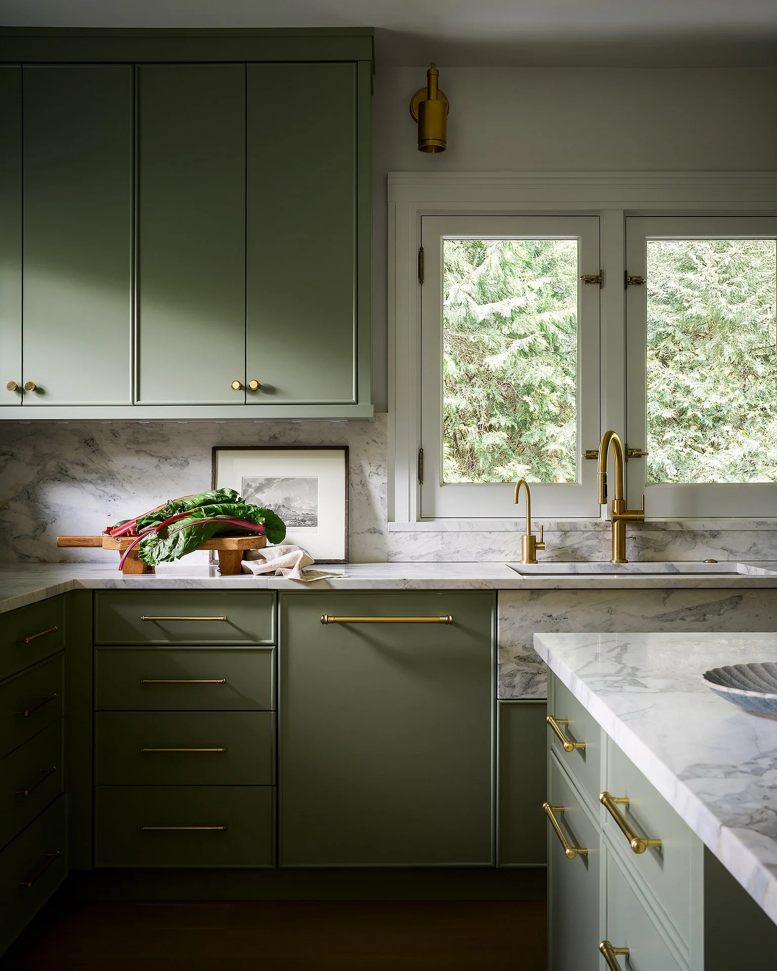 A modern kitchen with green cabinets, white marble countertops, and brass hardware. A window shows a view of trees outside, and a brass faucet is above the sink. There is a bunch of leafy greens on a cutting board on the counter.