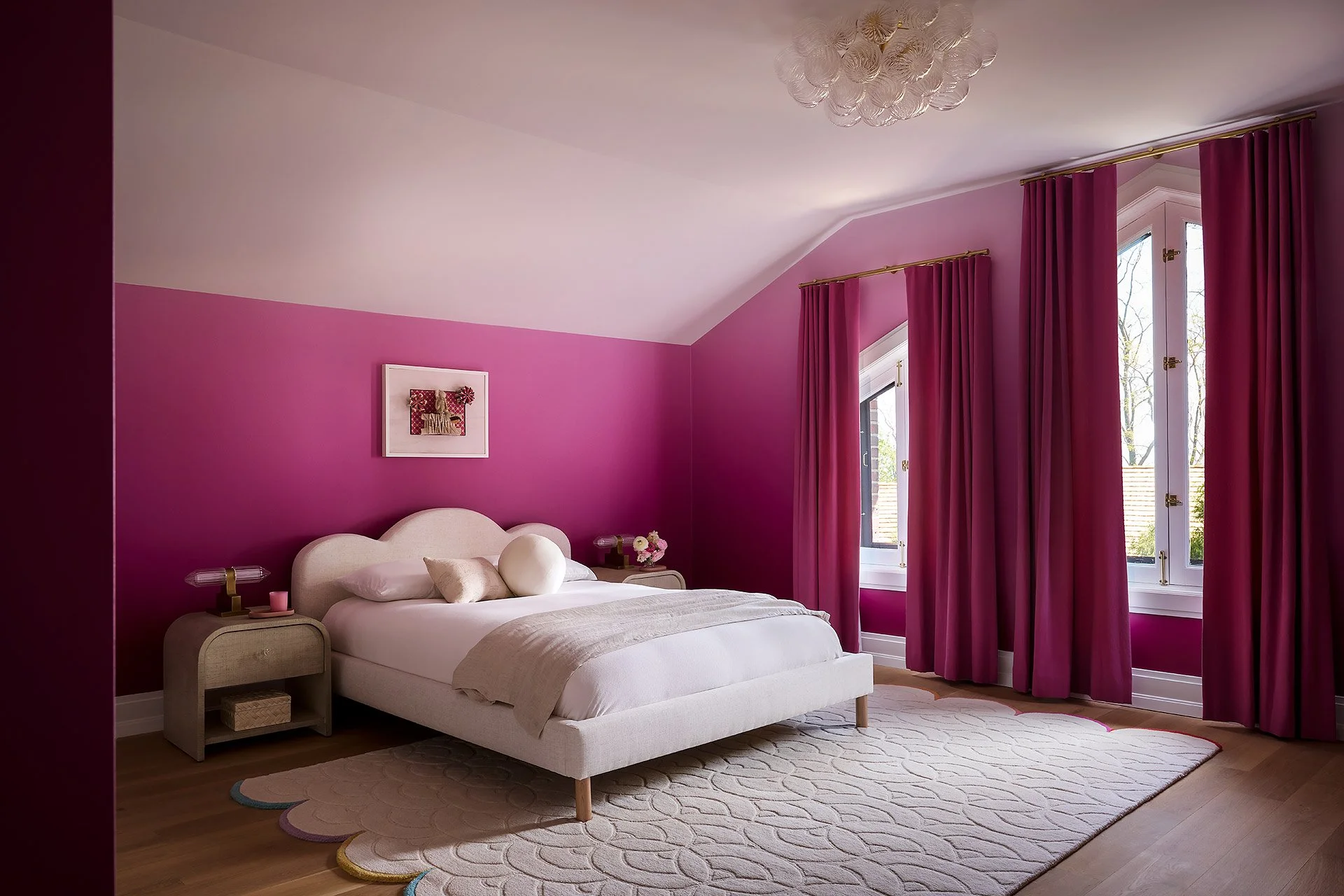 A bedroom with a pink accent wall, white bed with beige and pink pillows, nightstands with pink candles, framed artwork, large windows with pink curtains, and a modern glass chandelier.
