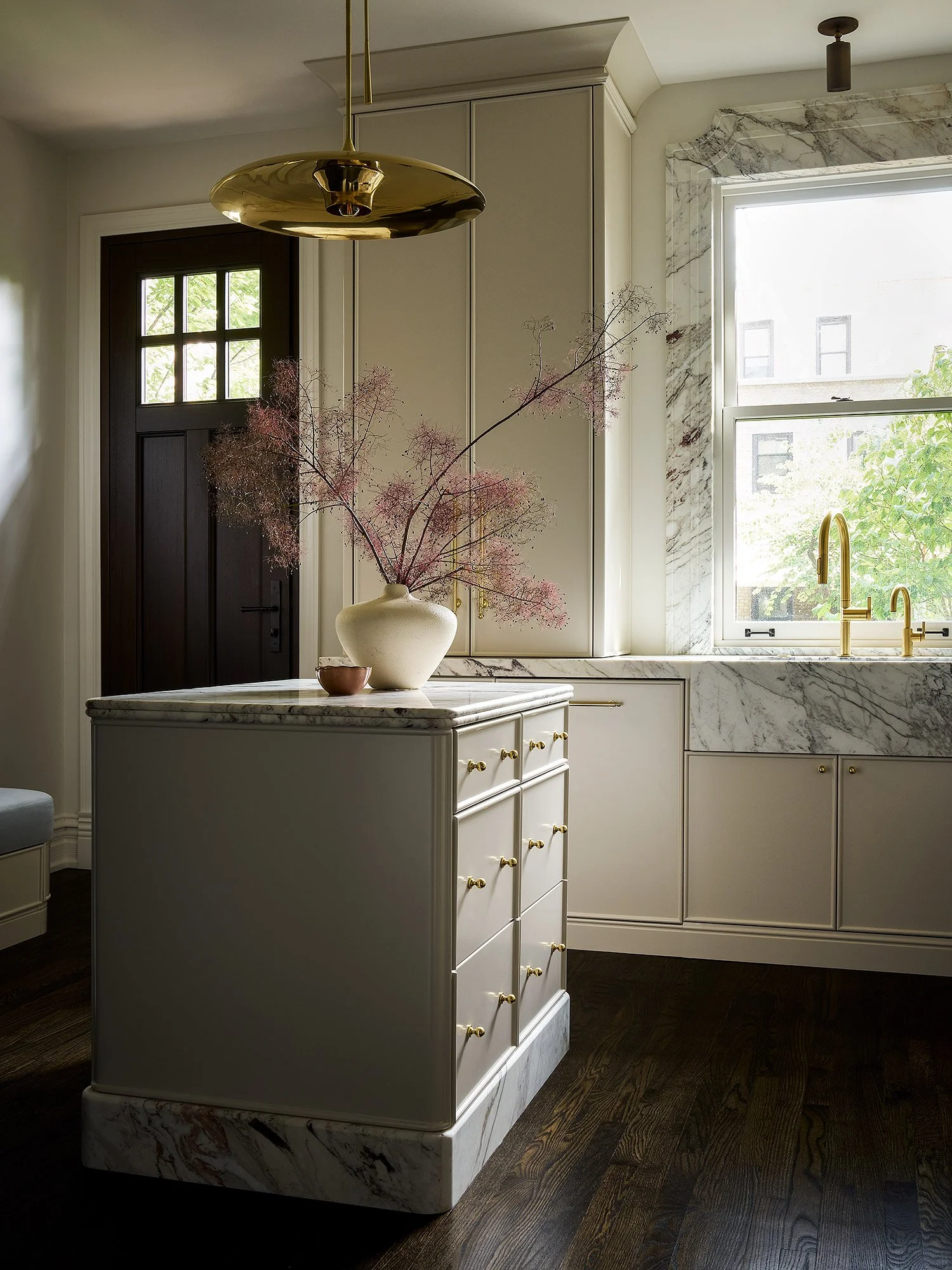 A kitchen with white cabinets, marble countertops, and a marble backsplash. There is a window with a view of greenery and a black door with four small windows. A brass faucet is visible on the countertop. A large white vase with pinkish branches is p