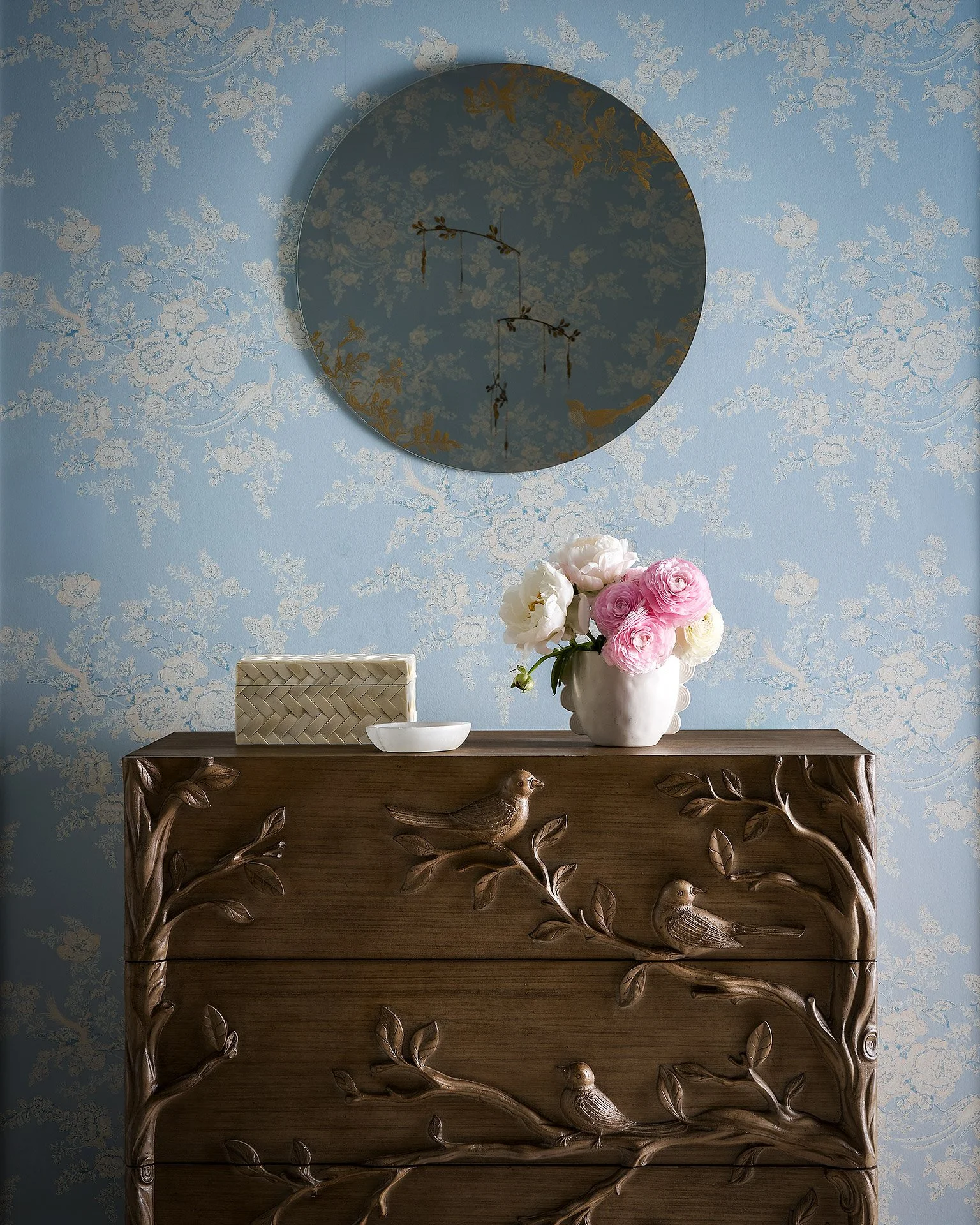 Wooden console table with carved birds and leaves design, topped with a white vase containing pink and white flowers, and a decorative box, against a blue floral wallpaper background with a round mirror reflecting the ceiling.