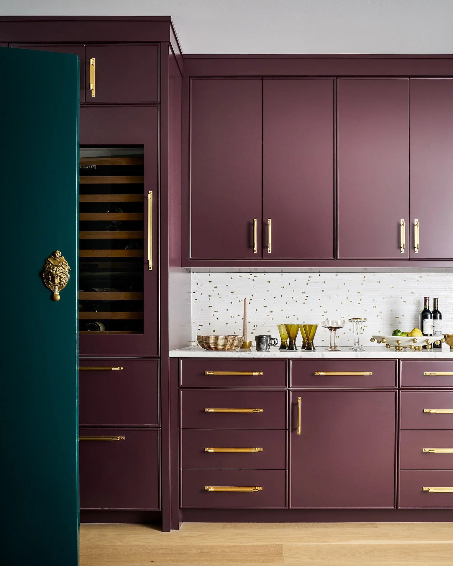 A section of a modern kitchen with purple cabinets, gold handles, and a white backsplash with gold accents. Part of a green door with a gold decorative handle is visible on the left.