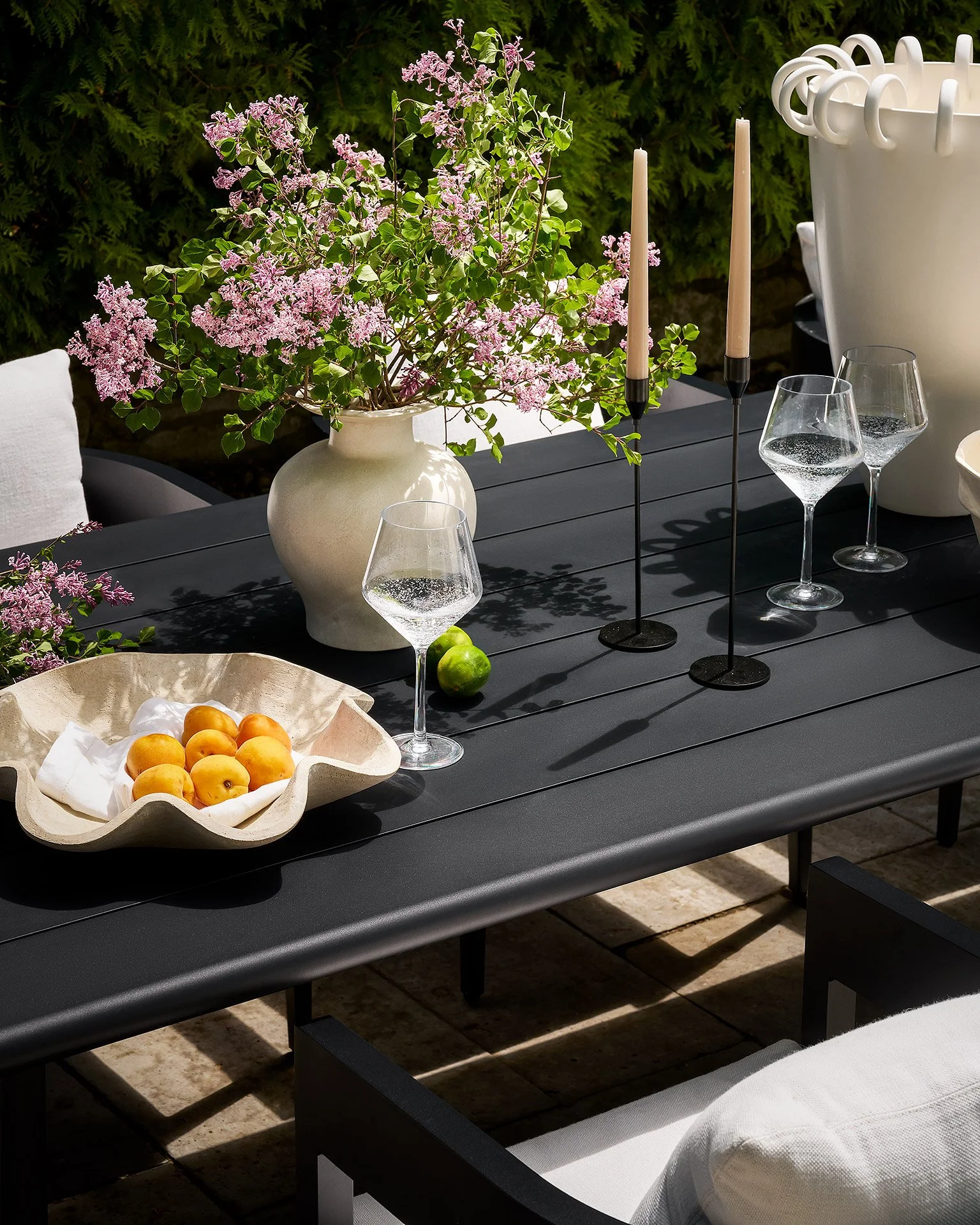 Outdoor dining table with a large white vase containing pink flowering plant, two pink candles, two glasses of water, a bowl of peaches, and some green apples, surrounded by greenery and white cushioned seating.