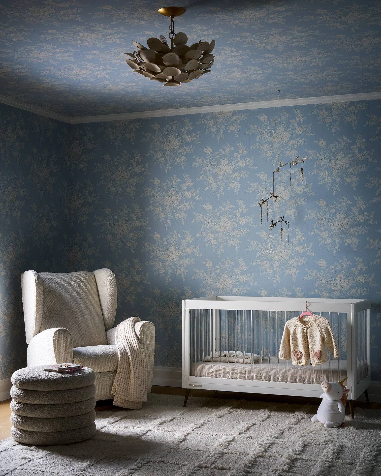 A nursery room with blue floral wallpaper, a white arched armchair with a blanket draped over it, a small stack of books on the armchair, a white crib with a plush bunny toy nearby, a hanging mobile, and a ceiling light fixture with numerous spoons a