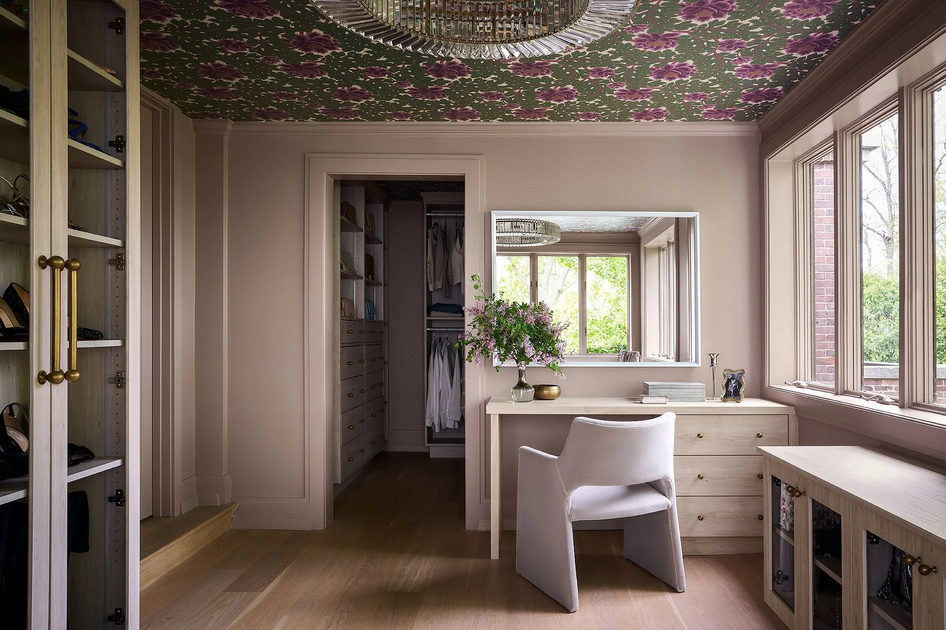 A home office with a floral patterned ceiling, large windows, a white desk, a chair, and decorative items like a vase with pink flowers and picture frames.