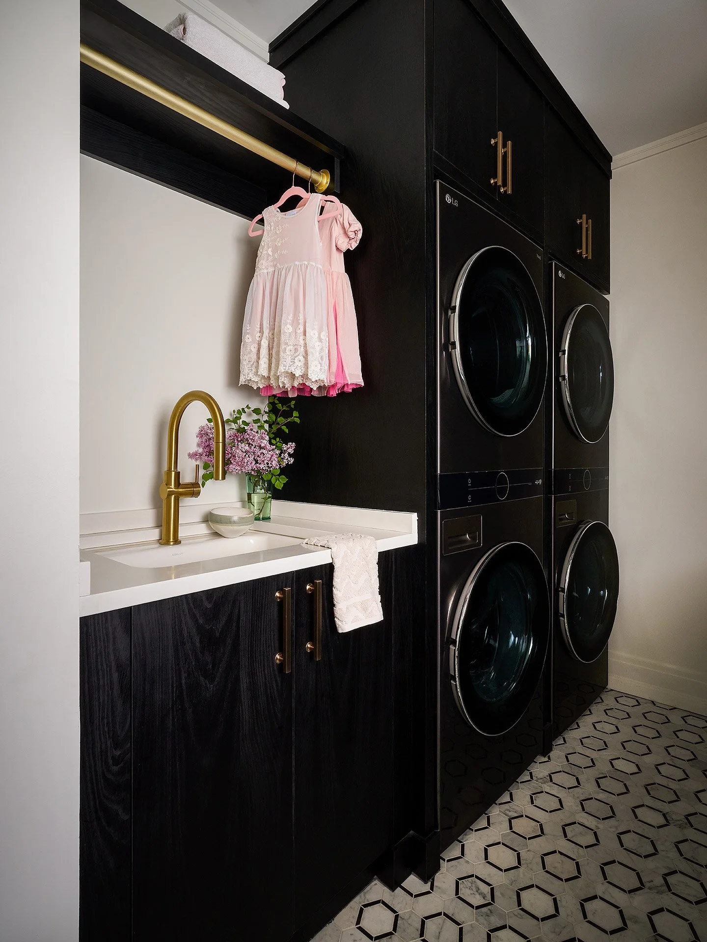 Laundry room with black cabinets, gold handles, front-loading washer and dryer, a small white sink with a gold faucet, a vase with pink flowers, a towel hanging on the side, and a clothes rod with pink and white dresses hanging.