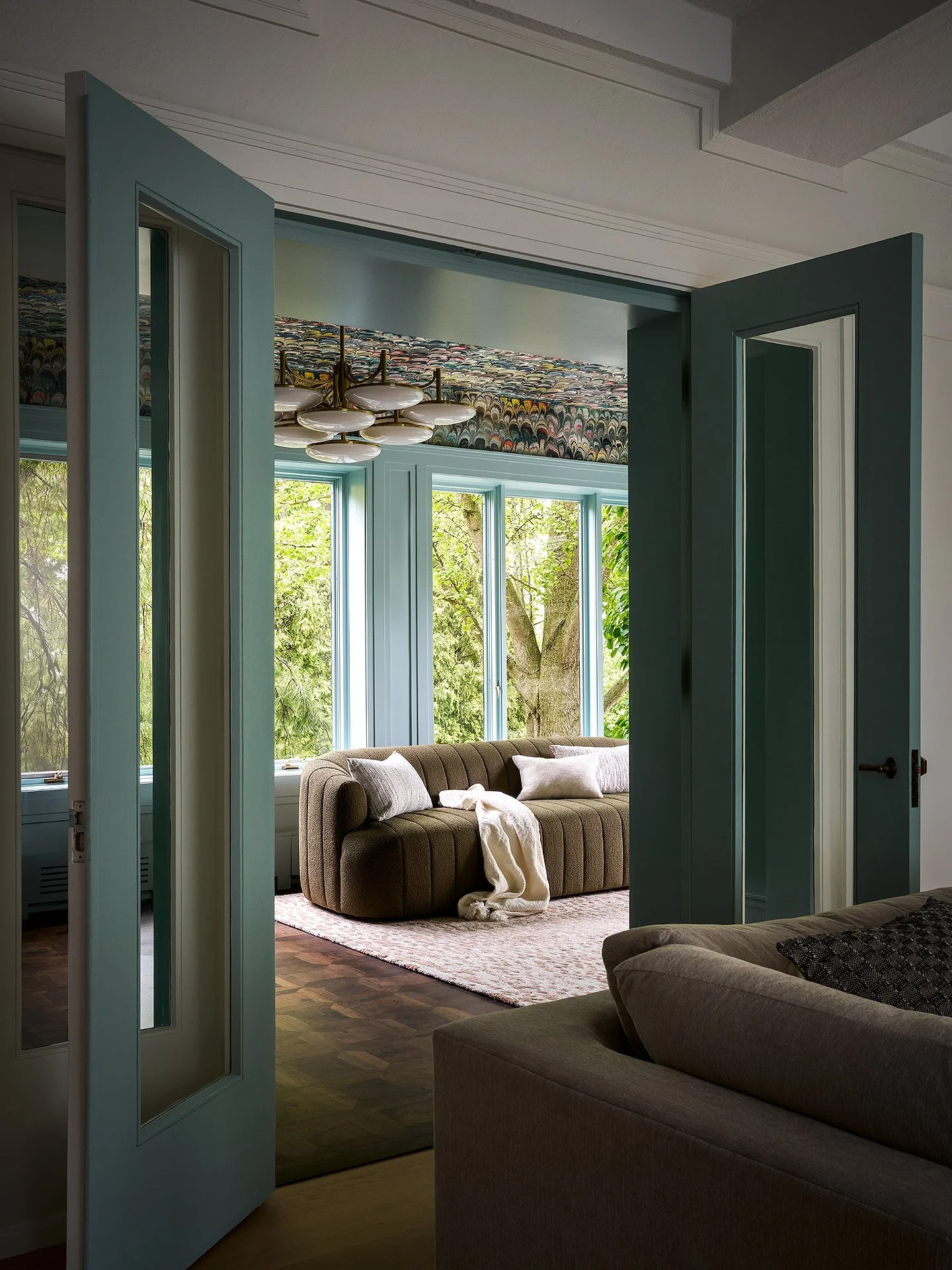 A living room viewed through open double doors with teal trim. The room has large windows showing green trees outside, a beige sofa with white pillows, a patterned wallpaper, and a modern chandelier.