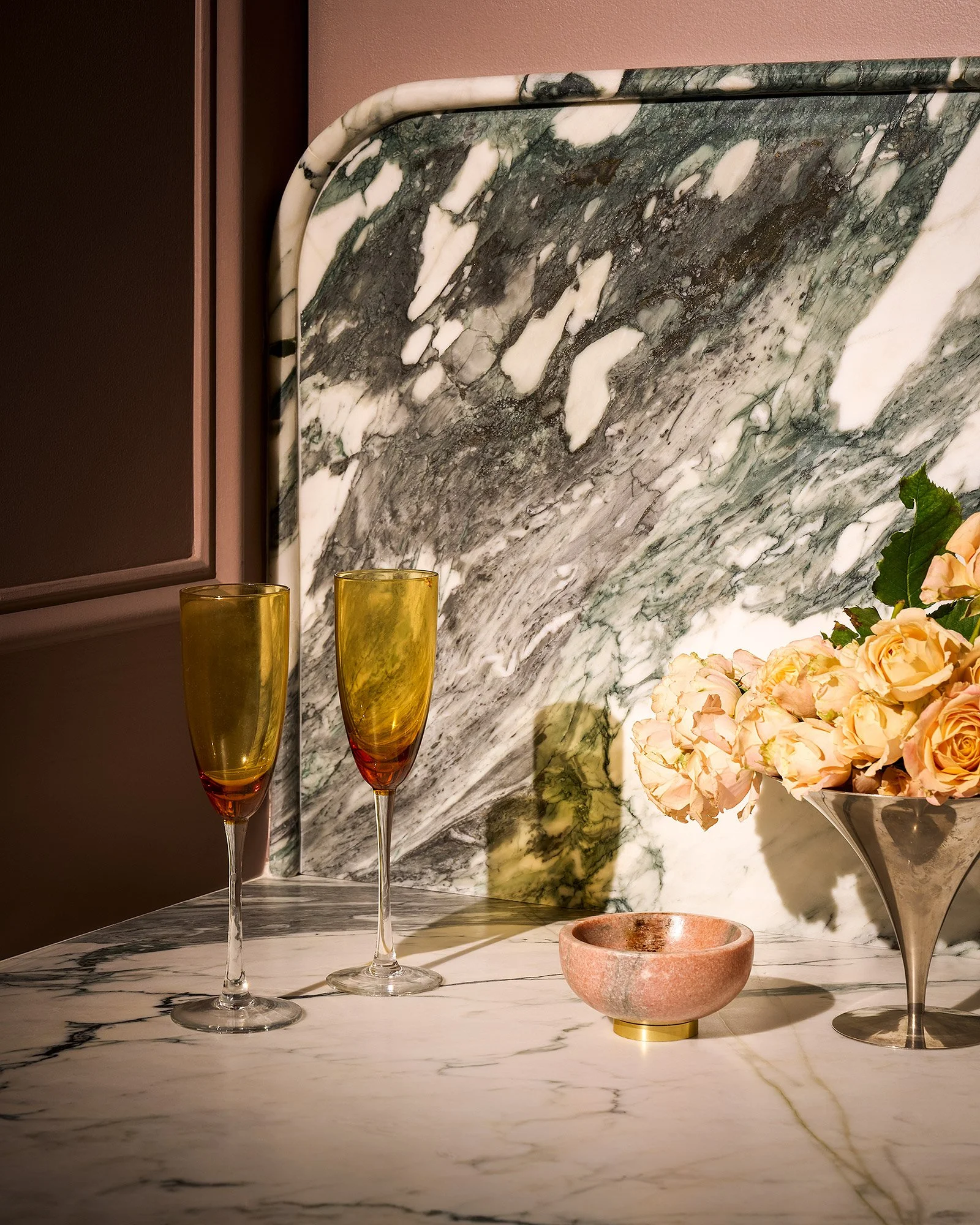 Close-up of a marble countertop with two tall champagne glasses, a small pink bowl, and a bouquet of light pink roses against a marble backsplash.