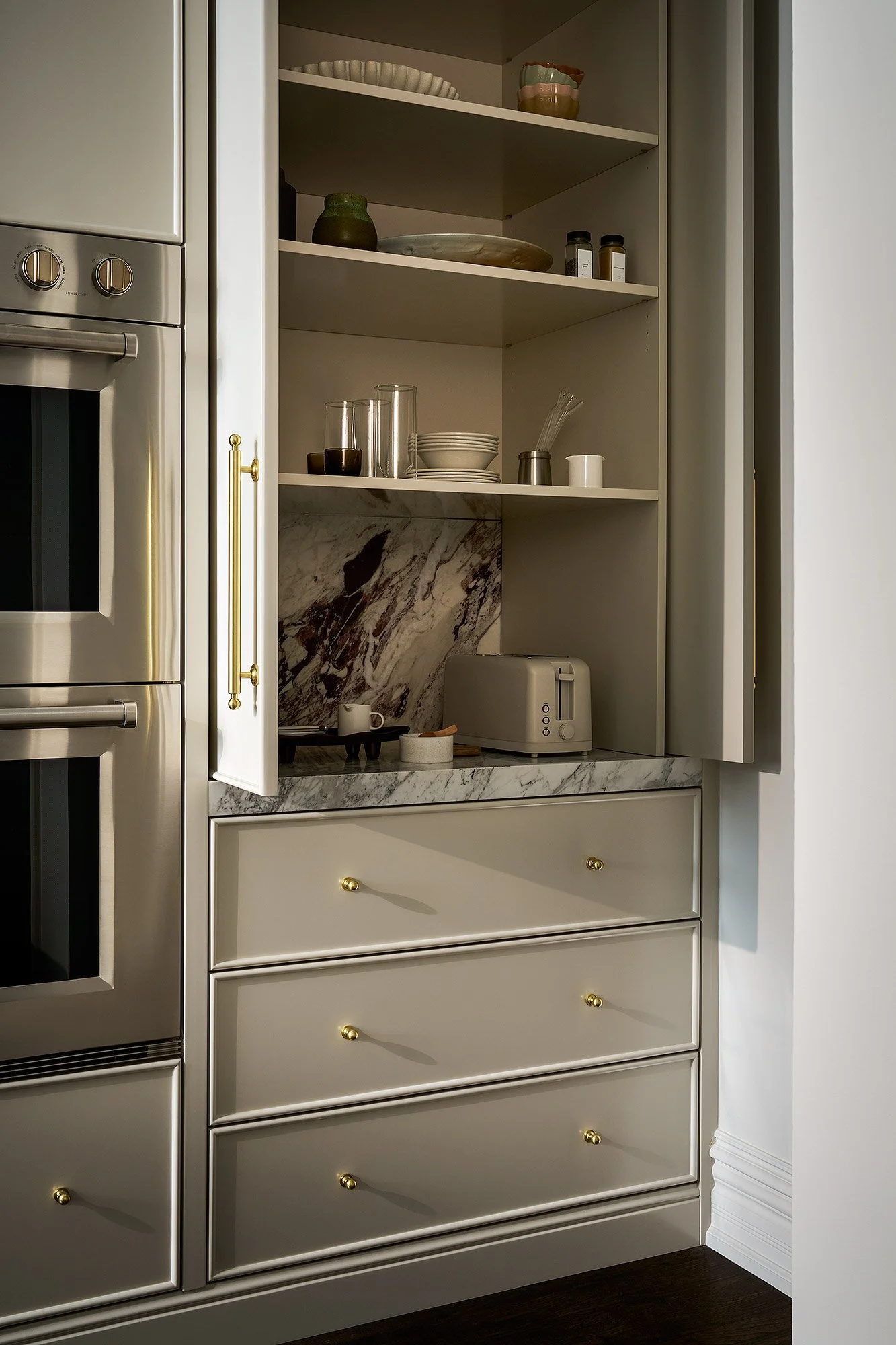 Open cabinet with shelves containing plates, bowls, glasses, jars, and kitchen utensils in a modern kitchen with marble countertop and white cabinetry with gold knobs.