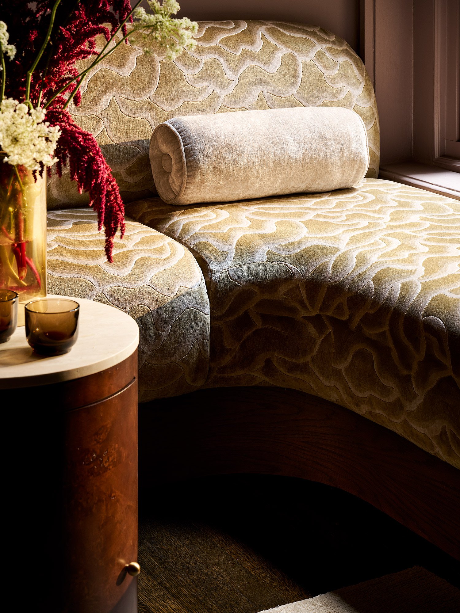 Close-up view of a cozy bedroom corner with a beige upholstered headboard, a cylindrical bolster pillow, a natural wood nightstand, a bouquet of colorful flowers in a glass vase, and a sunny window.