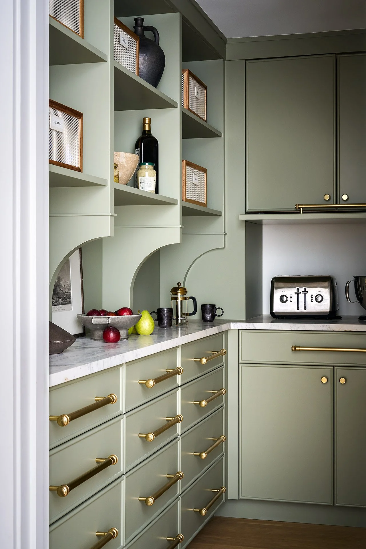 A kitchen with green cabinets, gold handles, open shelves with decorative items, a marble countertop with apples and pears, and a toaster on the counter.