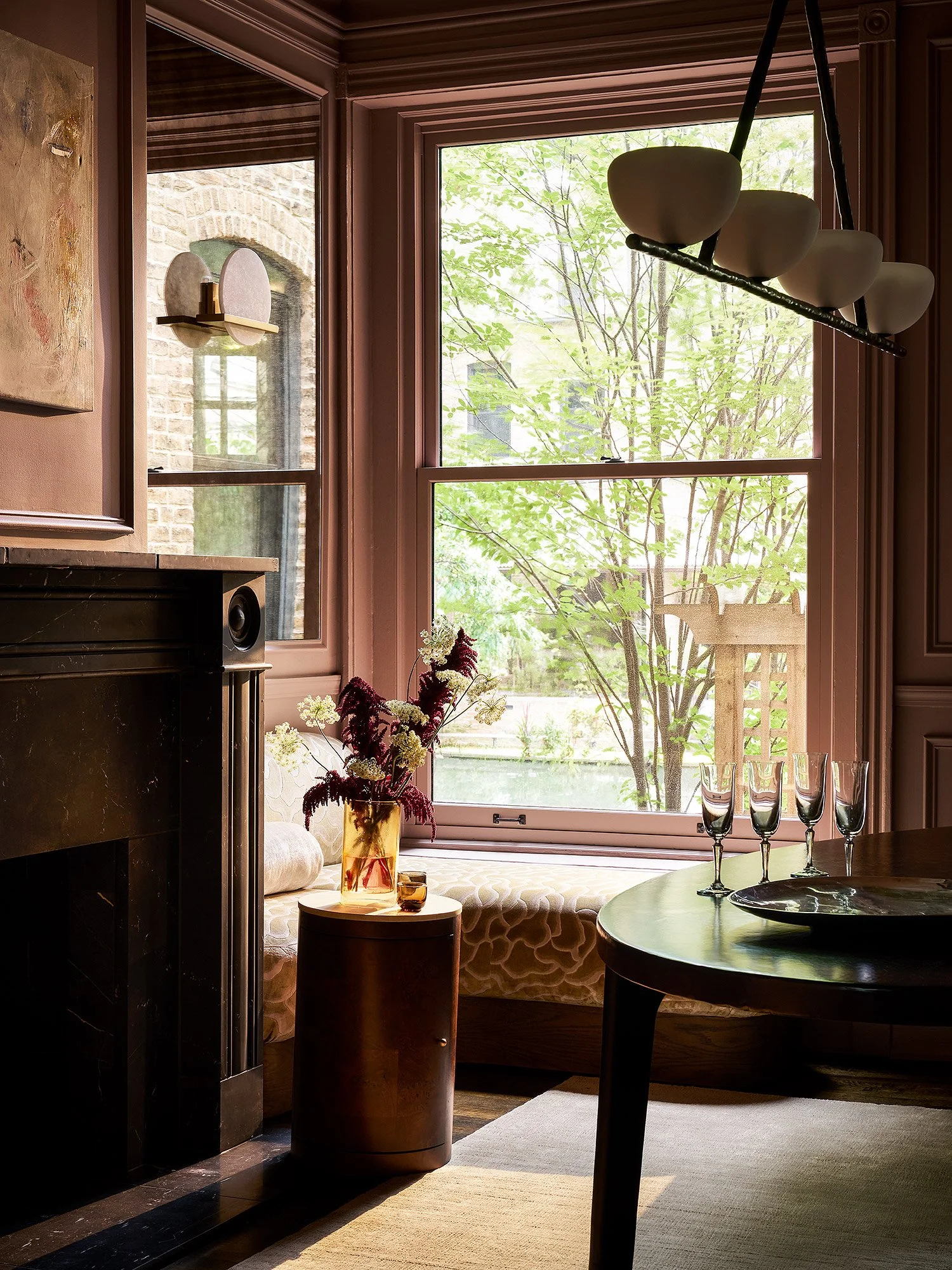 A cozy corner with a large window looking out onto trees, decorated with a flower arrangement, a small table, chairs, and a built-in window seat with cushions.