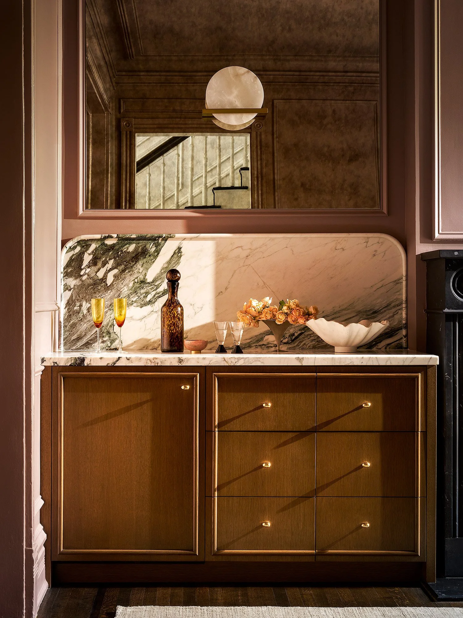 A kitchen interior with a marble countertop, decorative bottles, glasses, and a flower arrangement, featuring a large mirror and wooden cabinets.