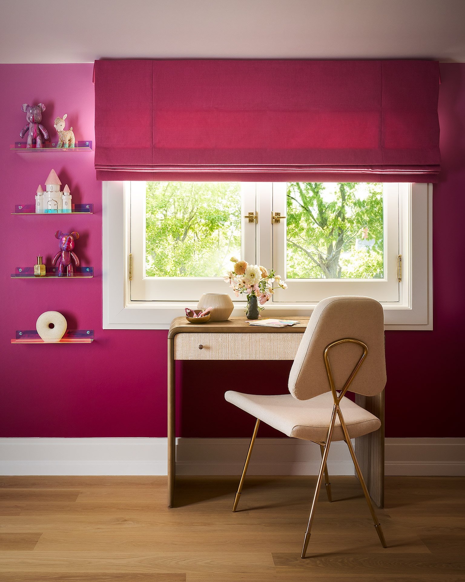 A cozy workspace with a pink wall, a window with greenery outside, a white desk with a vase of flowers, a beige chair with gold legs, and pink shelves with stuffed animals and decorative items.