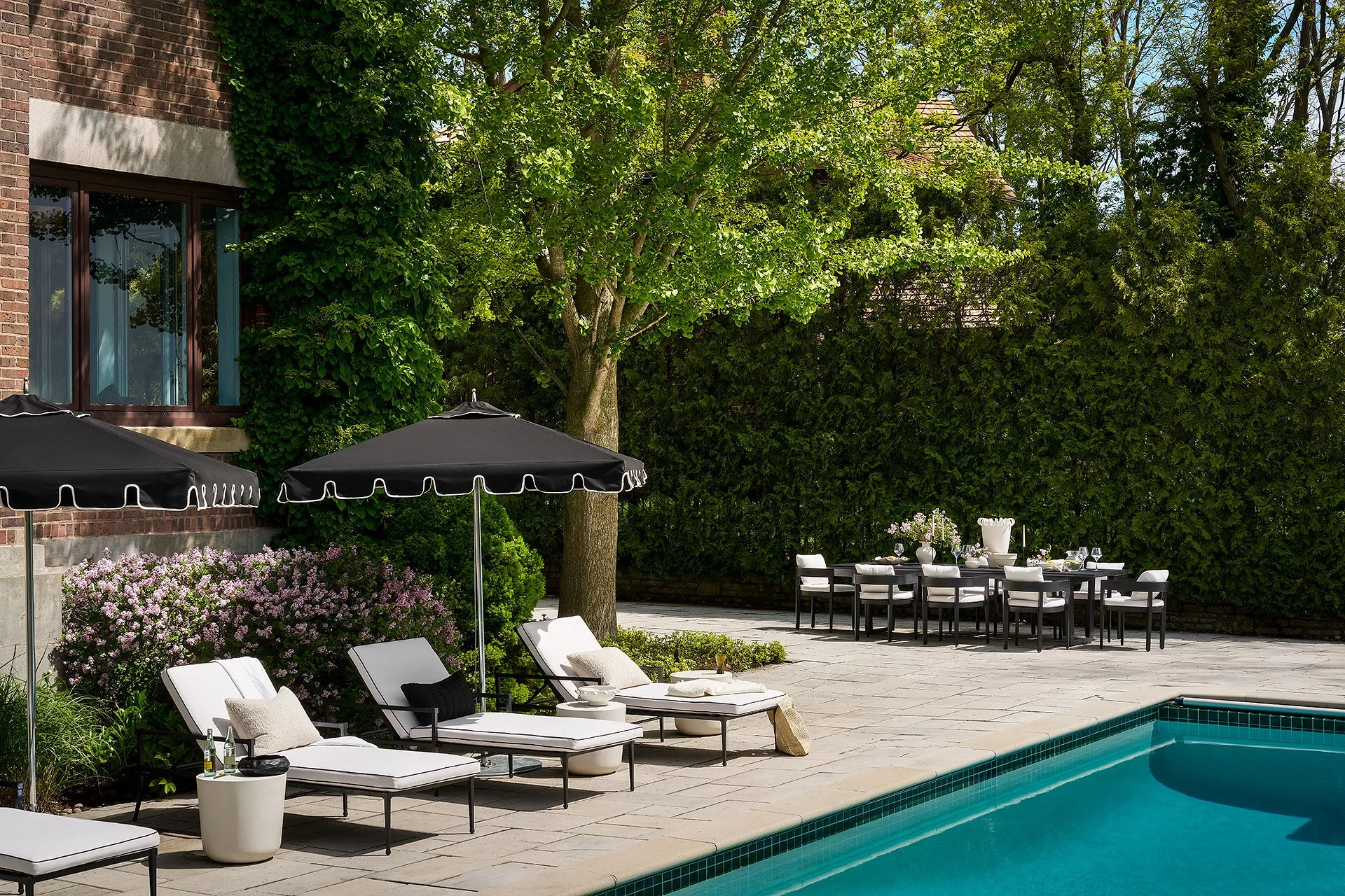 Outdoor backyard with swimming pool, lounge chairs, patio table with chairs, umbrellas, lush greenery, and a brick house wall.