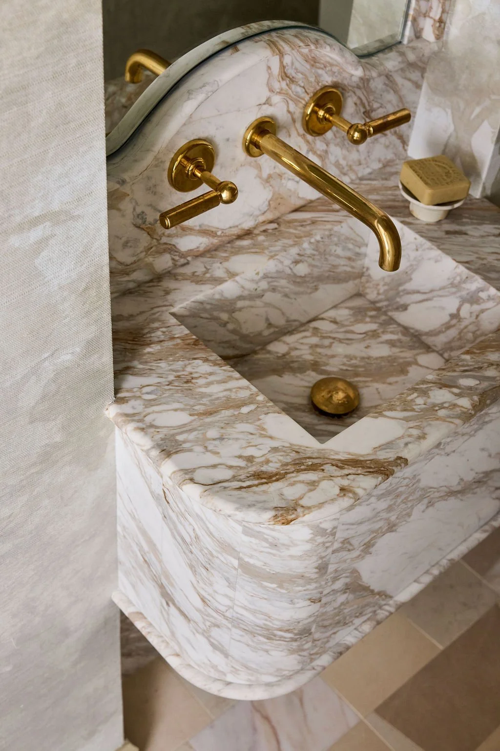 Marble sink with gold-colored faucet and handles, a bar of soap on a dish, and a mirror reflected on the wall.