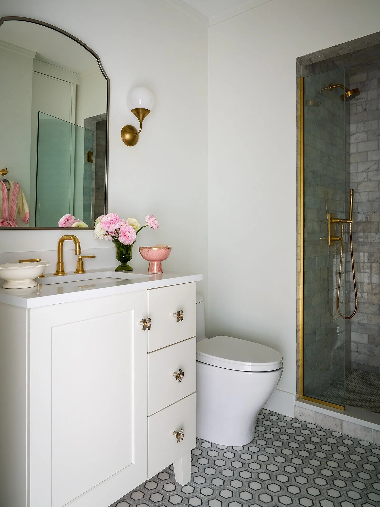 Modern bathroom with white vanity, gold fixtures, pink flowers in a green vase, a pink bowl, toilet, and a walk-in shower with marble tiles and gold accents.