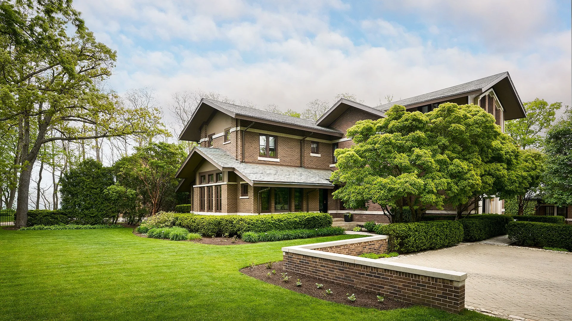 A large multi-story brick house with a landscaped front yard, green grass, trees, bushes, and a paved driveway under a partly cloudy sky.