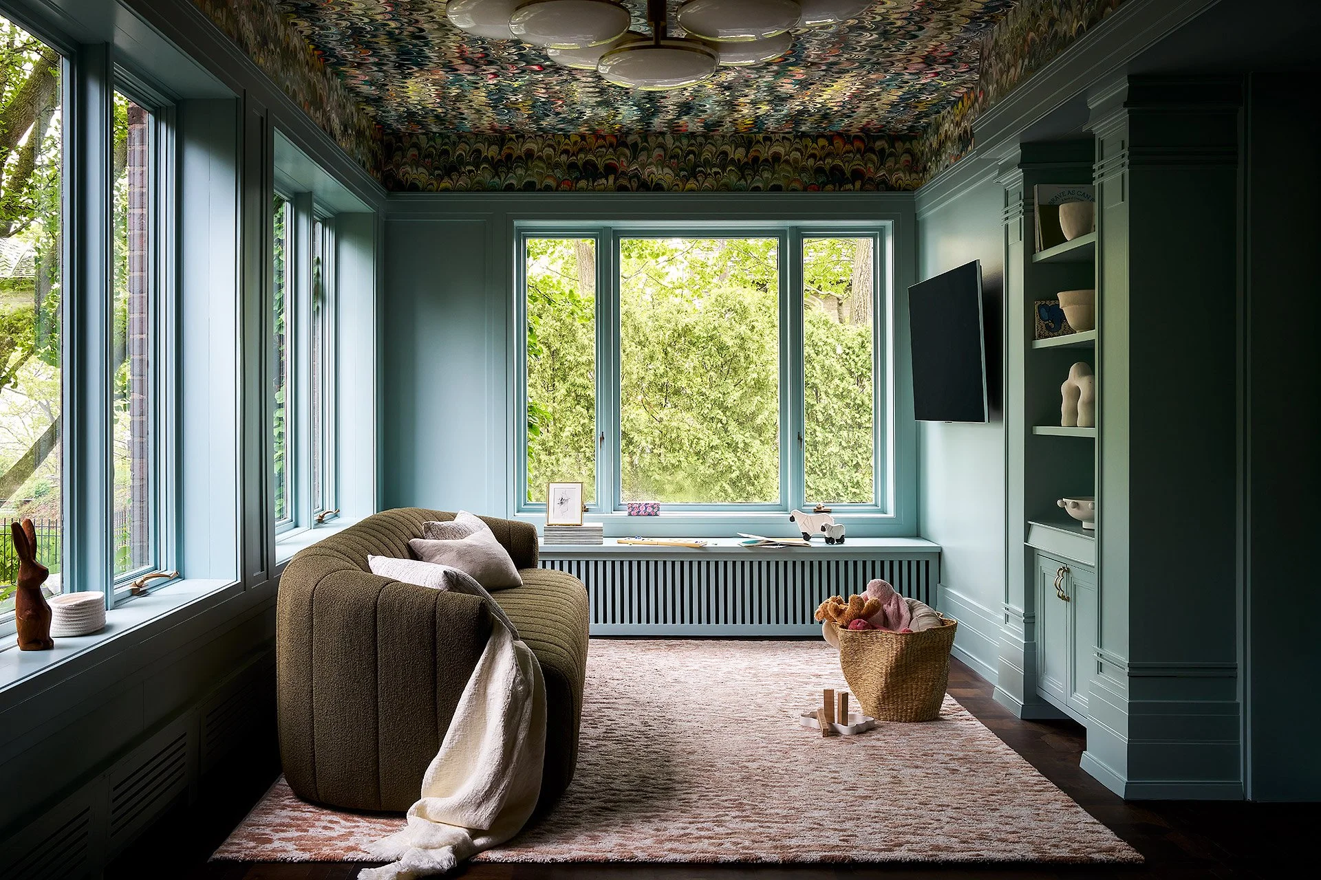 Living room with large windows showing green trees, blue walls, built-in shelves with decorative items, a beige sofa with pillows, and toys in a basket, illuminated by natural light.