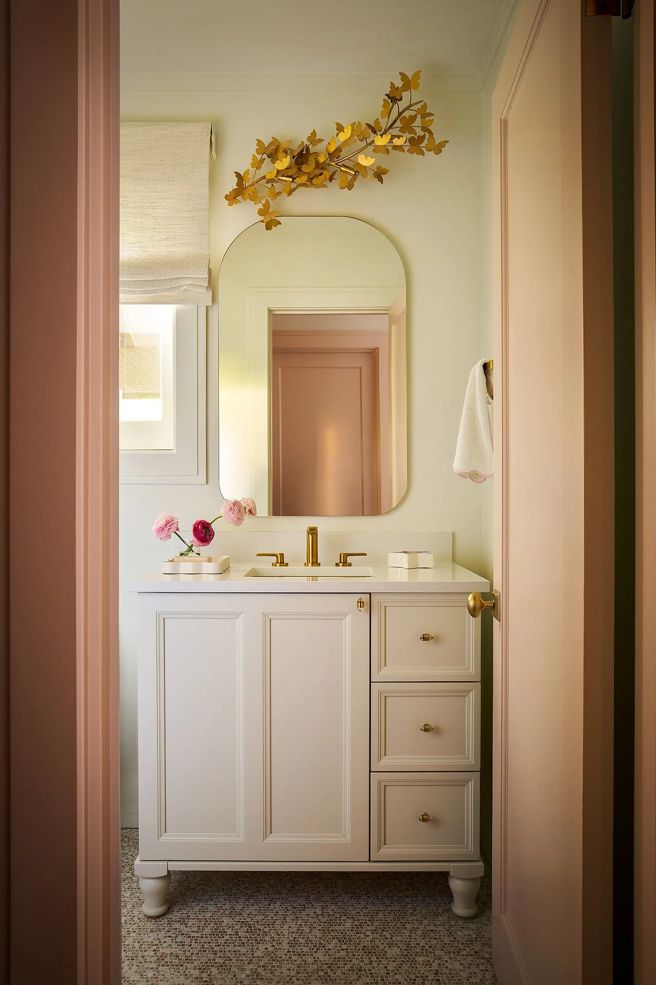 A small white bathroom vanity with gold fixtures and a mirror above, decorated with pink flowers in a white vase and a small box, with a peach-colored wall and a window with a beige Roman shade.