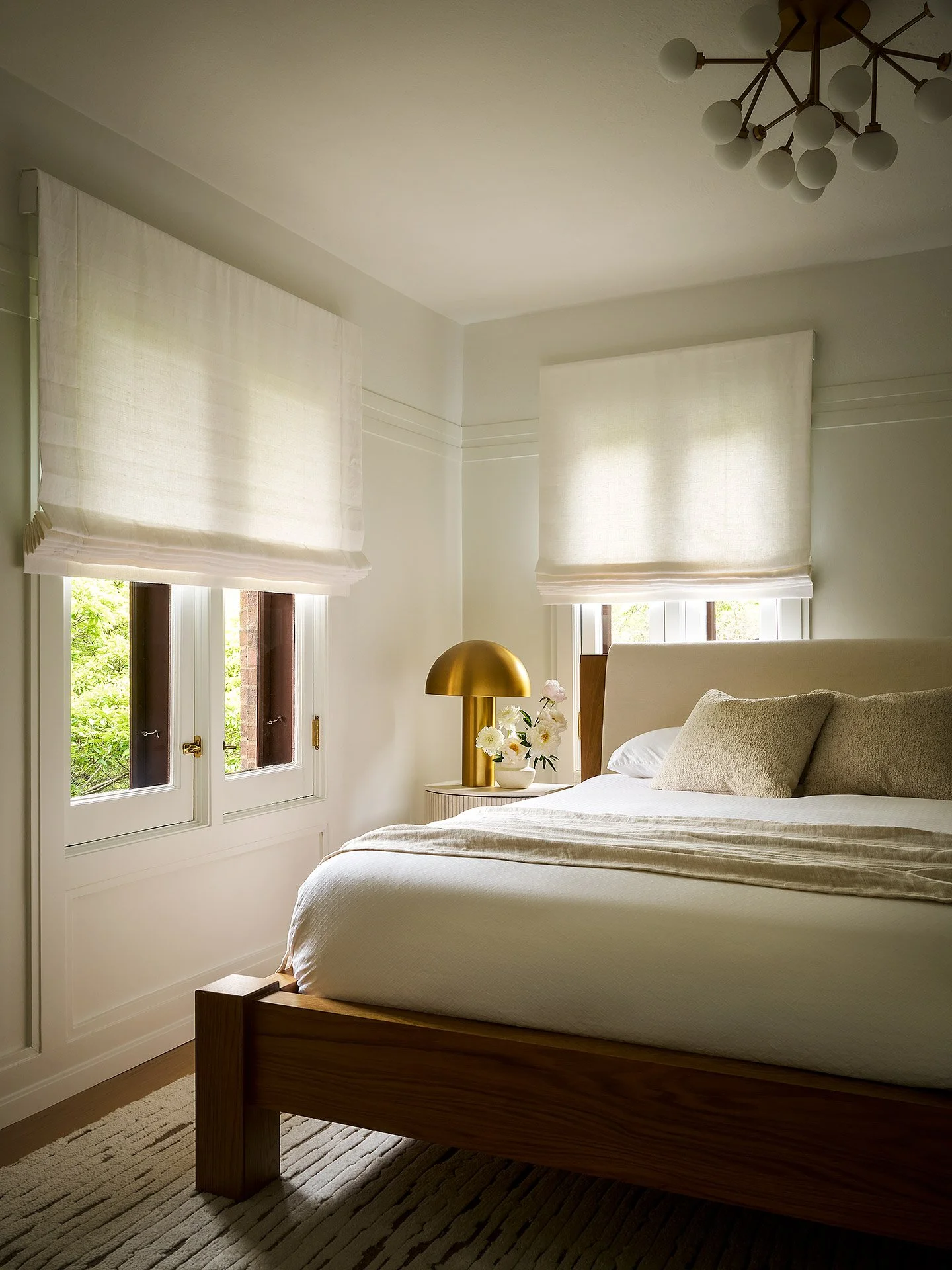 A cozy, well-lit bedroom with a large bed, white bedding, and beige pillows. There are two windows with white Roman shades, a gold bedside lamp, and a vase of white flowers on a small table. A modern chandelier hangs from the ceiling.