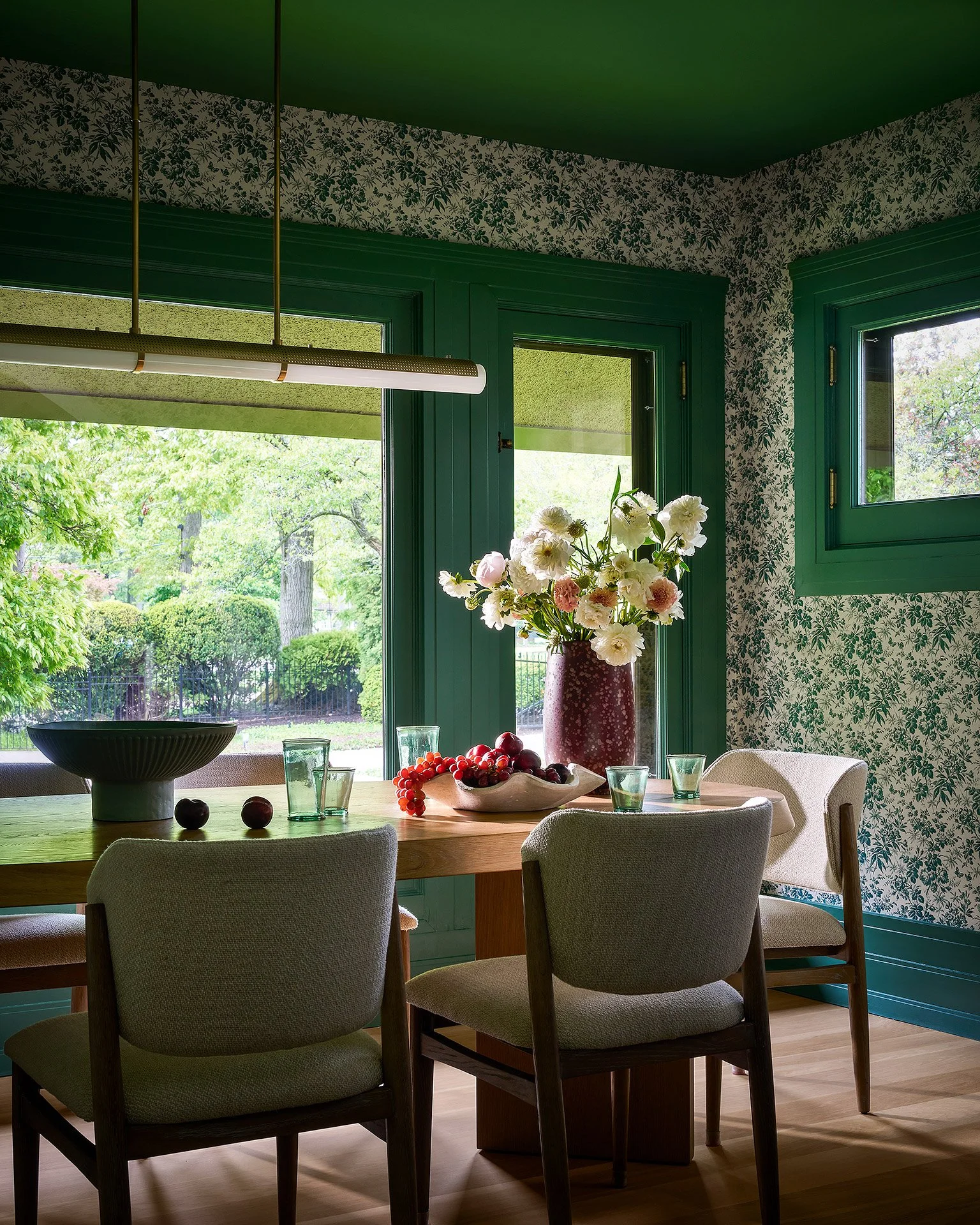 A dining room with a wooden table, beige upholstered chairs, and a large window with green trim. On the table is a white bowl with grapes, a white vase with white and pink flowers, four glasses, and a few cherries and plums. The walls have green and 