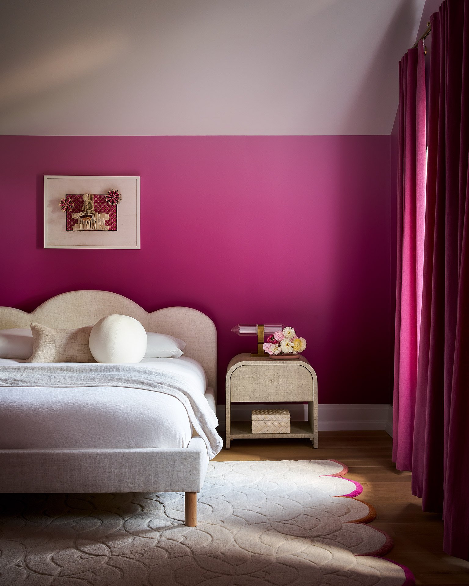 A bedroom with a white bed, beige headboard, and pillows. The wall behind the bed is painted pink with a white upper section. There is a nightstand with a pink and white lamp and a small bouquet of flowers. Pink curtains are on the window, and a fram